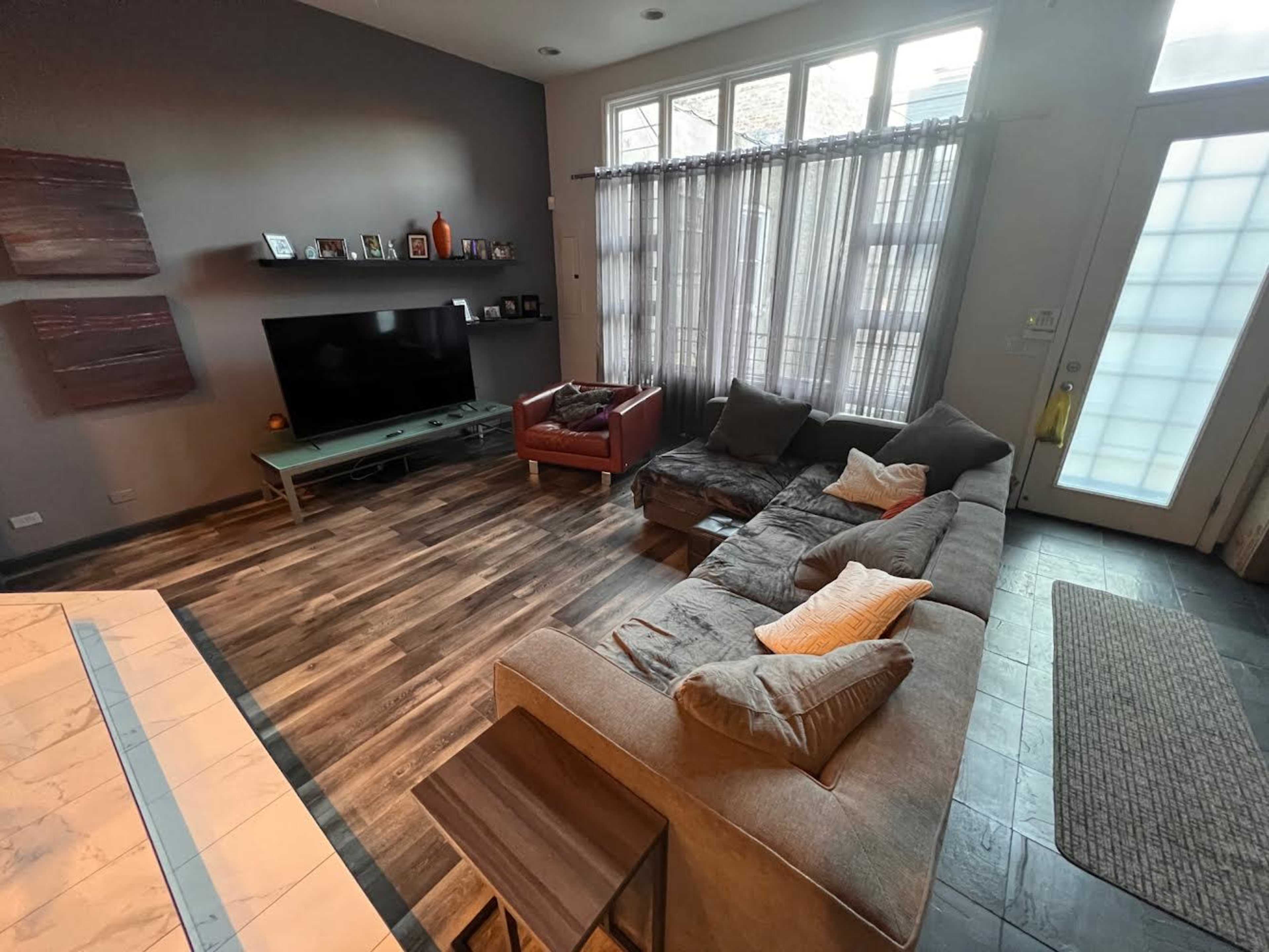 A living room with a large sectional sofa, a television on a light green stand, and a red armchair, illuminated by natural light coming through tall windows.