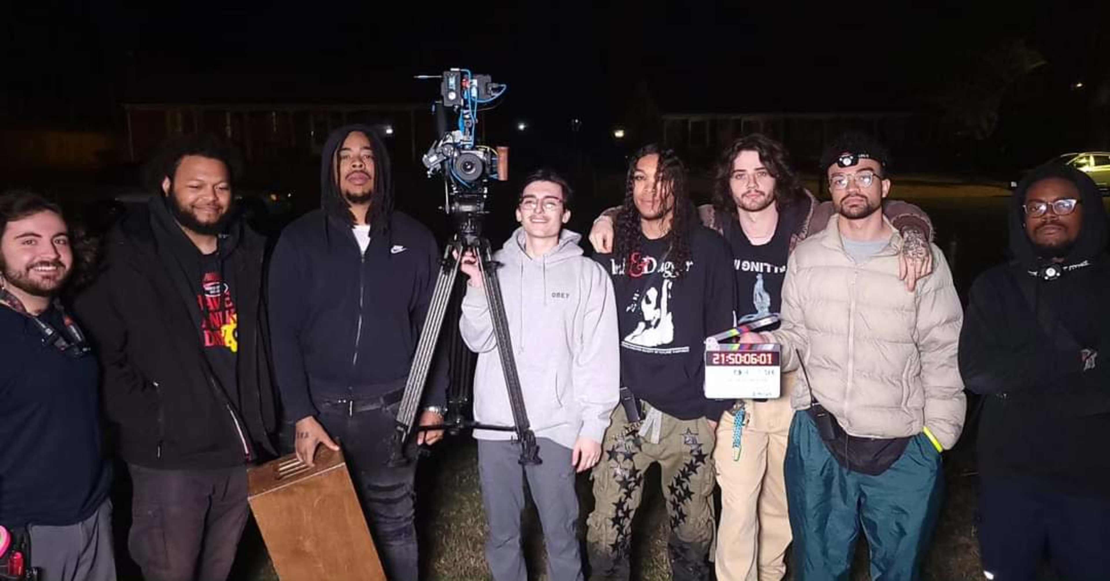 A group of eight individuals poses together outdoors at night, with one person holding a camera on a tripod.