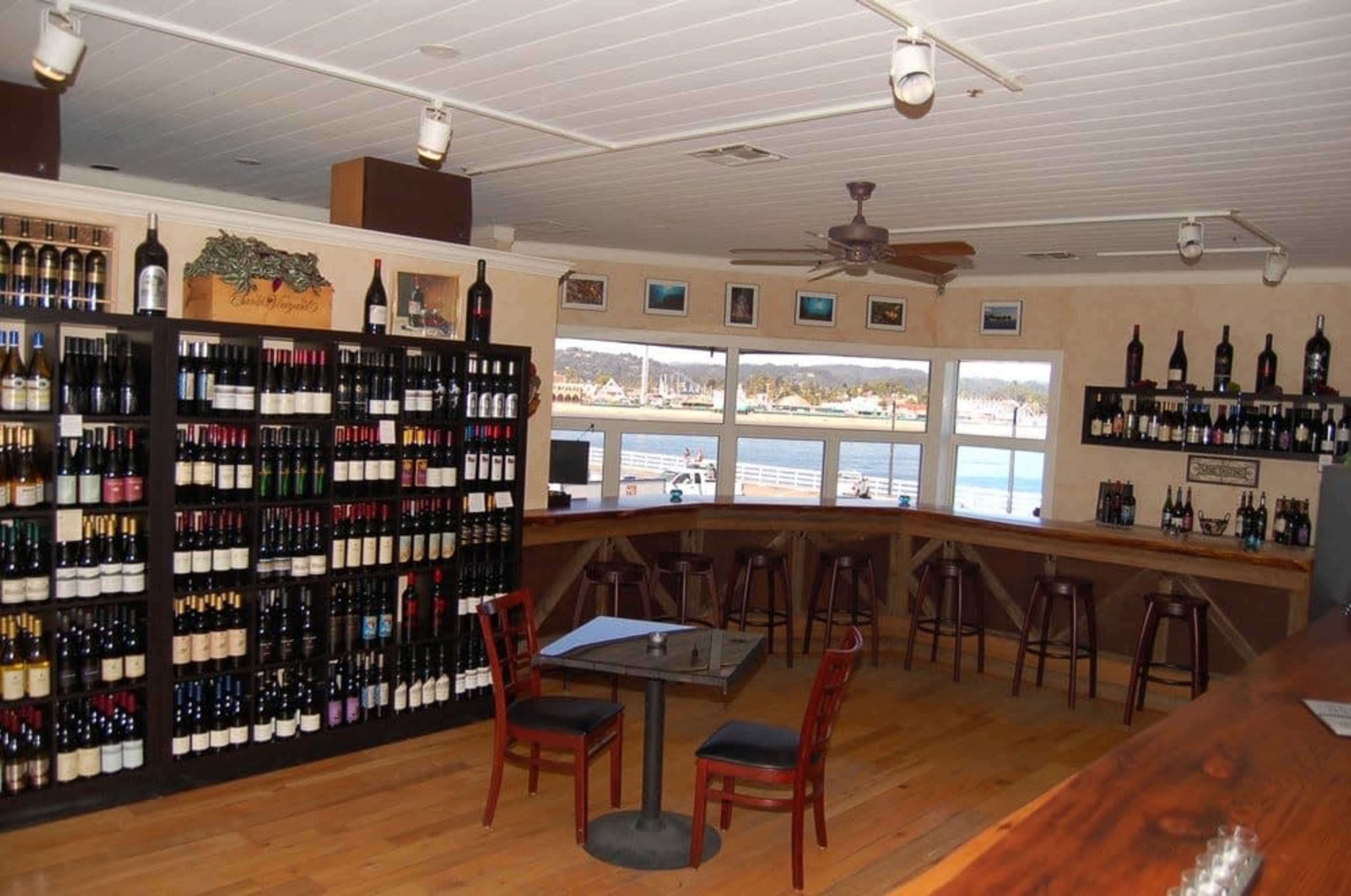 The image shows a wine shop interior featuring rows of wine bottles on shelves and a small seating area with a table and chairs near large windows.
