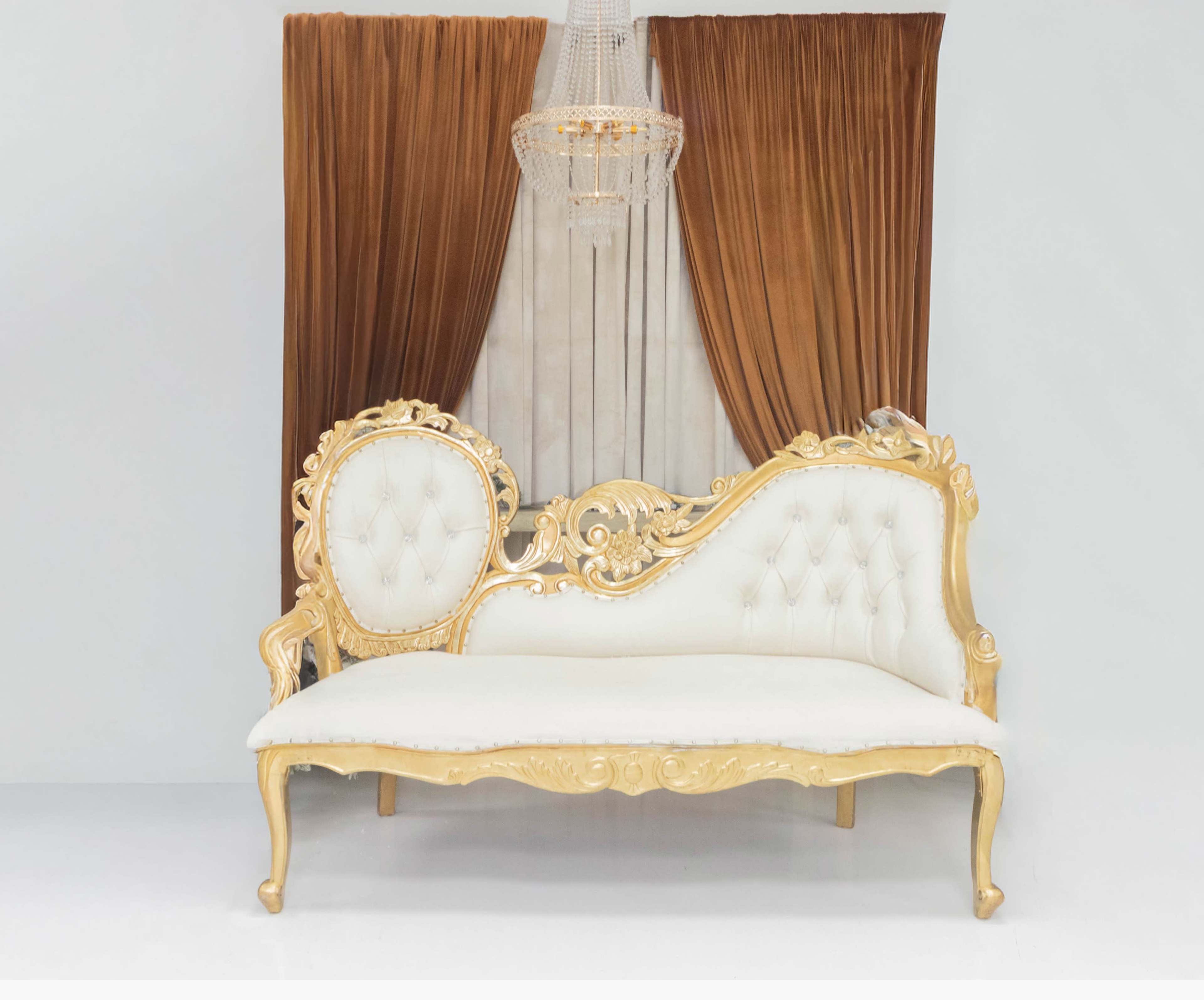 The image shows an elegant white and gold upholstered bench with intricate detailing, positioned in front of rich, brown drapes and a crystal chandelier.