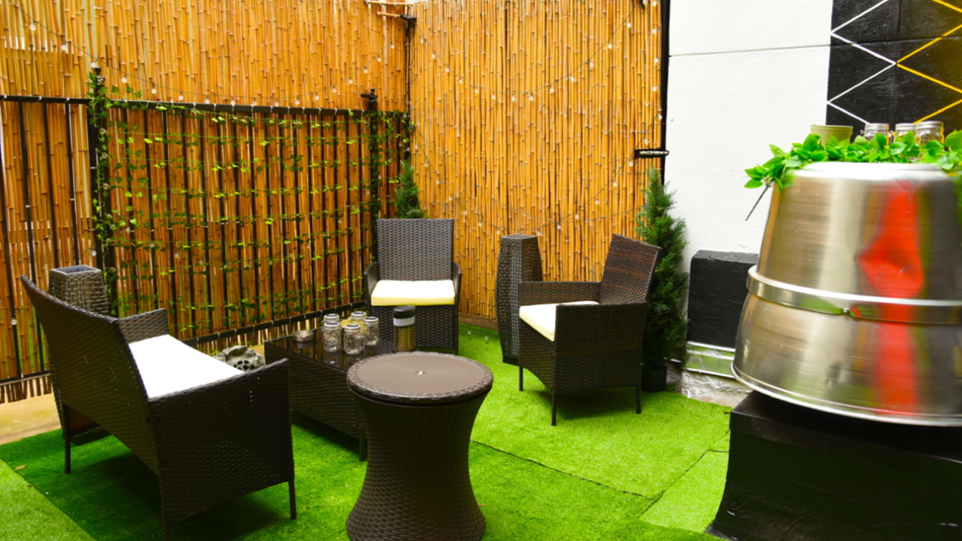 A small outdoor seating area with wicker chairs, a circular table, and a metal container, surrounded by bamboo fencing and artificial grass.
