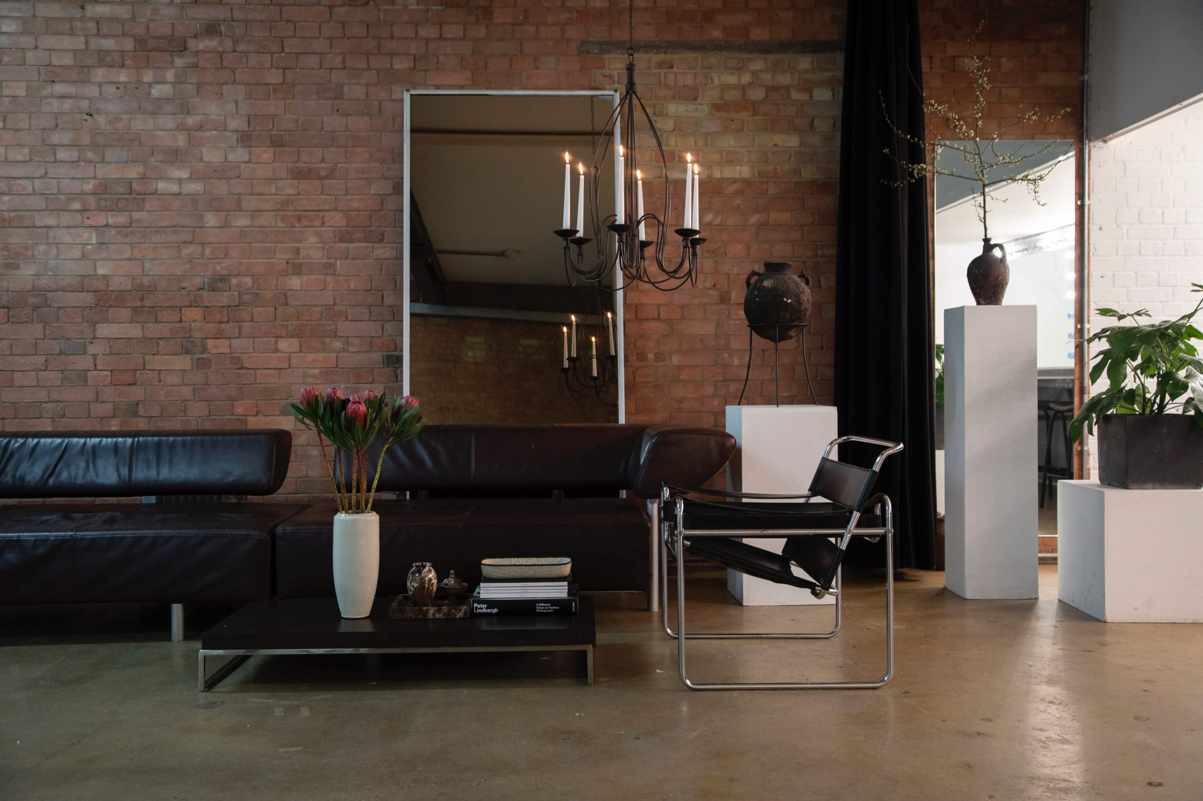 A modern living room features a dark leather sectional sofa, a glass coffee table, and a large mirror reflecting a chandelier and decorative elements against a brick wall.