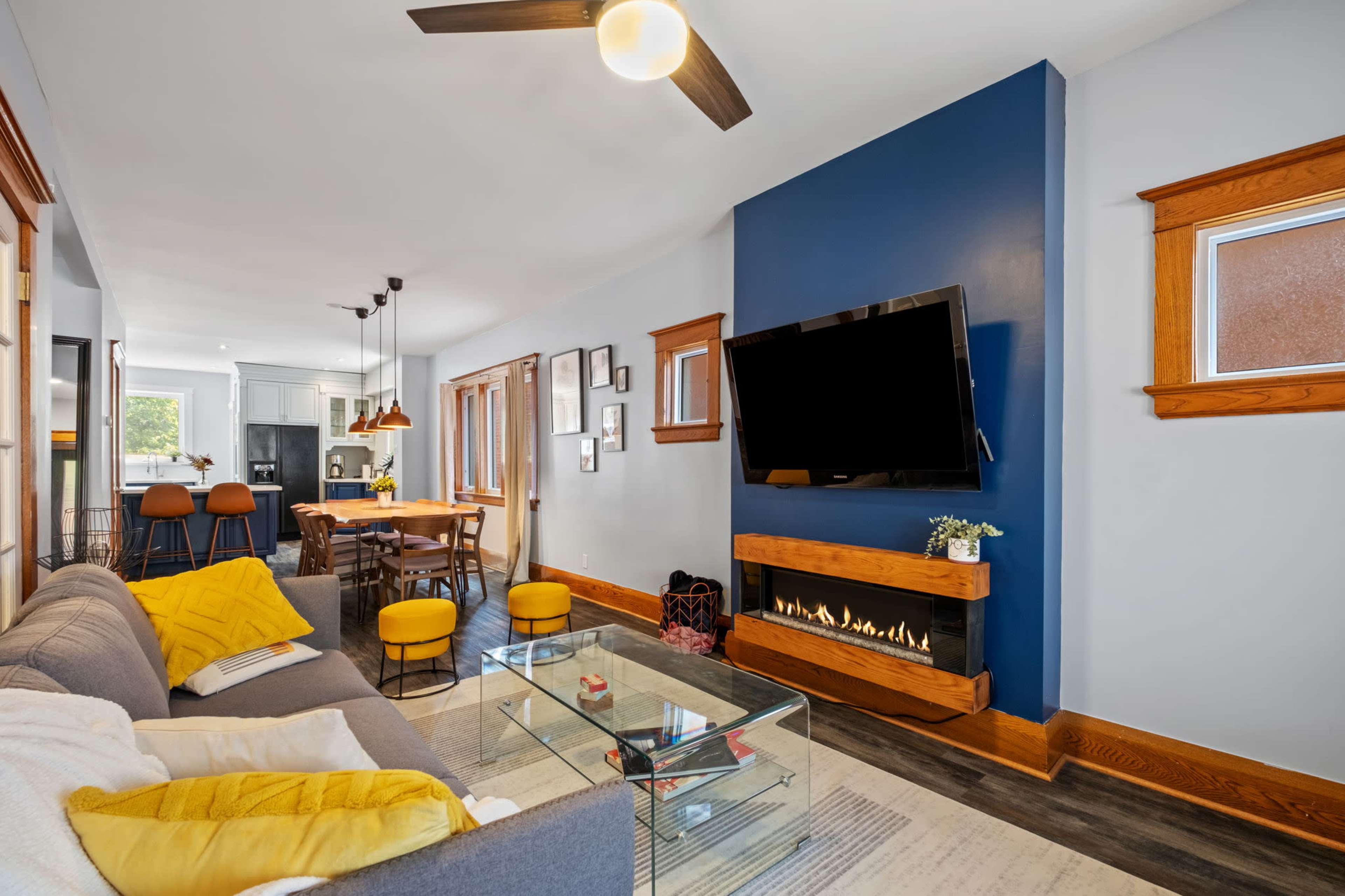 The living room features a modern design with a gray sofa, a glass coffee table, a wall-mounted television, and a decorative electric fireplace, complemented by yellow accent pillows and barstools nearby.