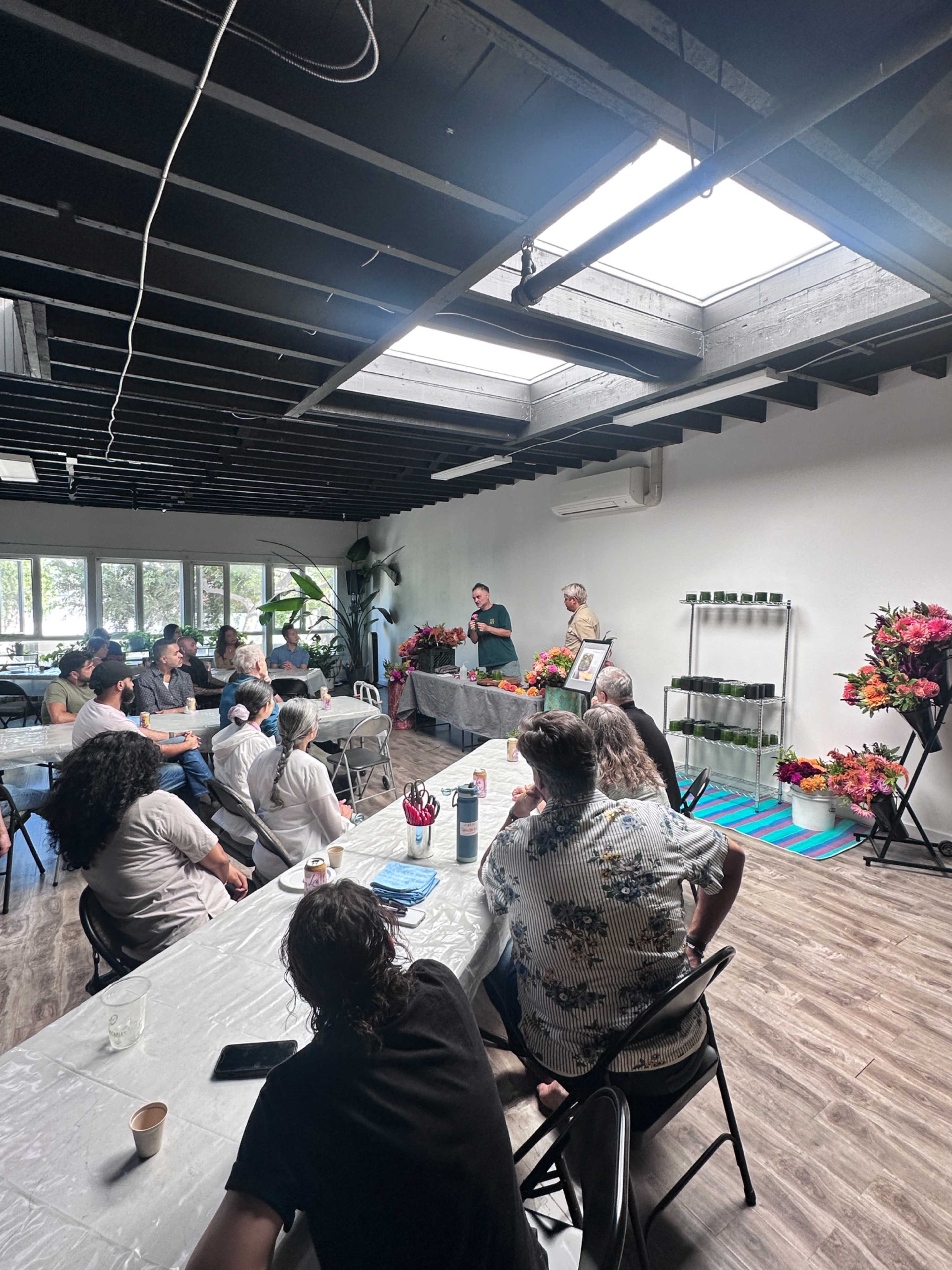 A group of people sits at tables in a spacious room while a presenter stands at the front, displaying materials surrounded by colorful floral arrangements.