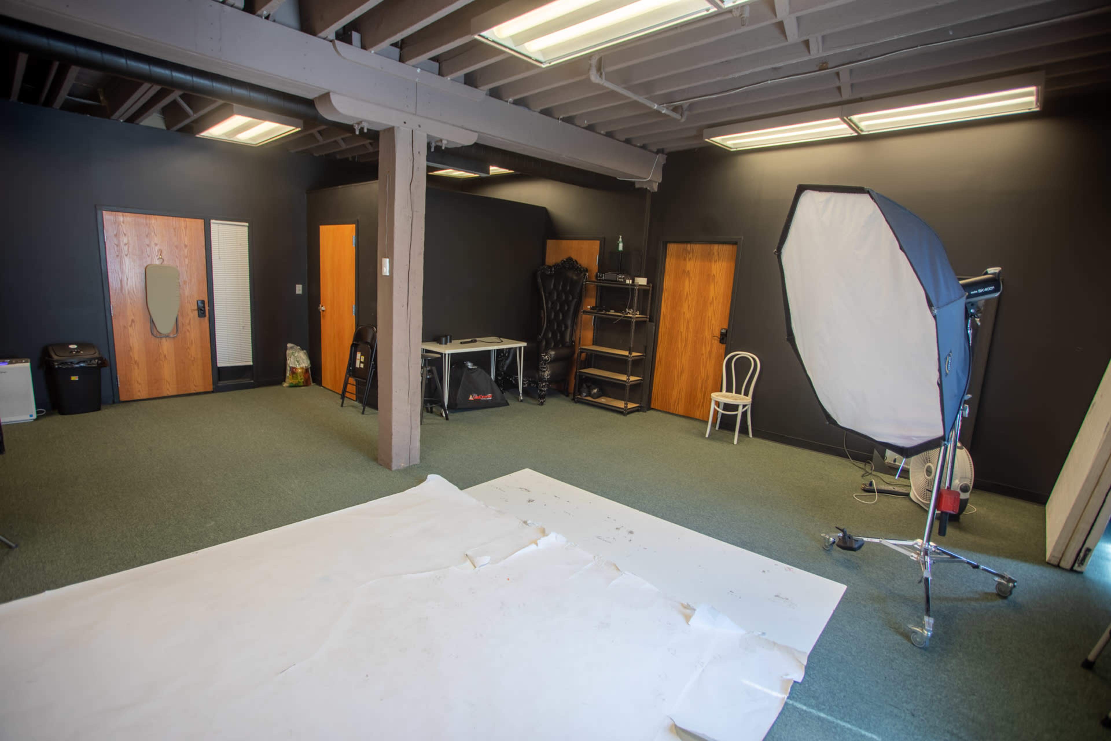 A photography studio with a large softbox light, a white backdrop on the floor, and several wooden doors against dark walls.