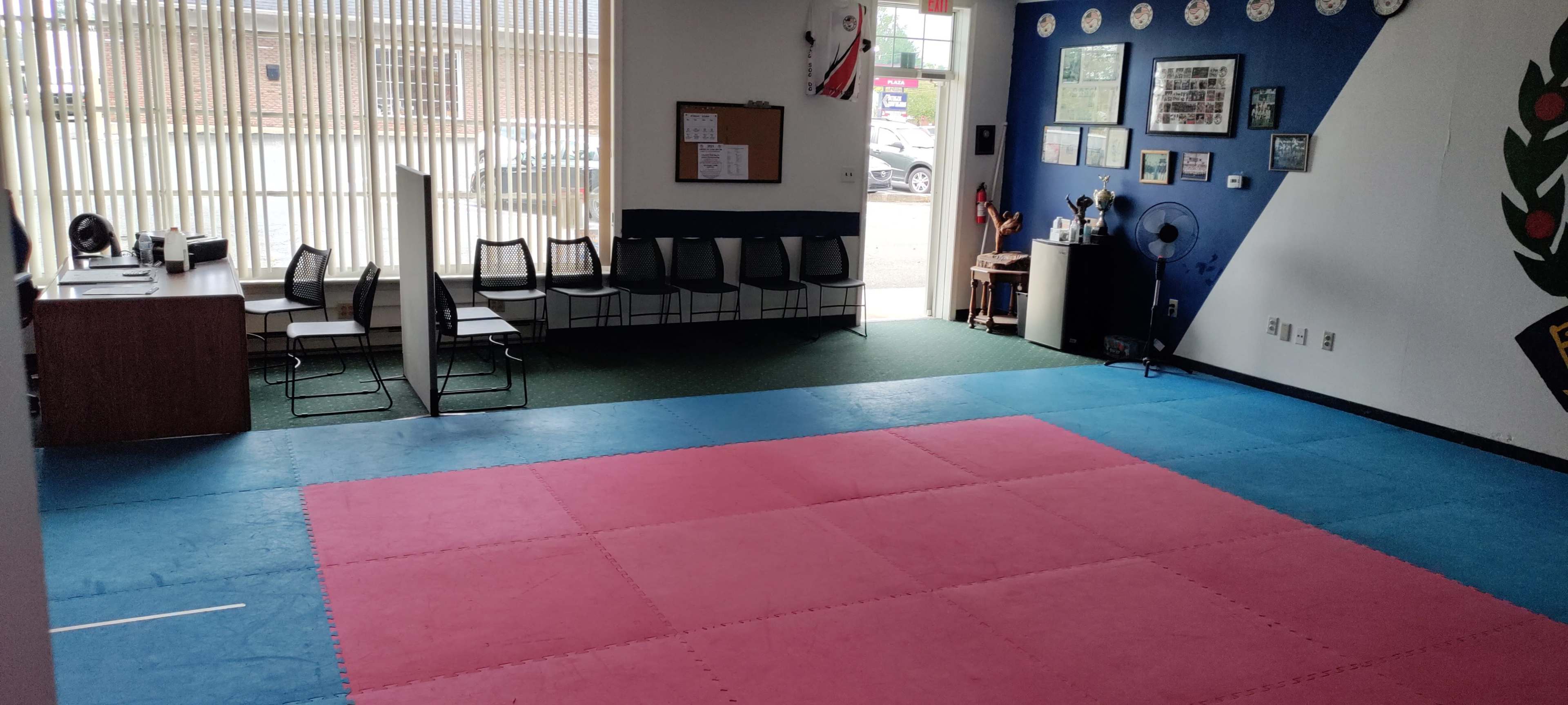 A martial arts studio with a pink and blue mat area, lined chairs, and a reception desk against the wall.