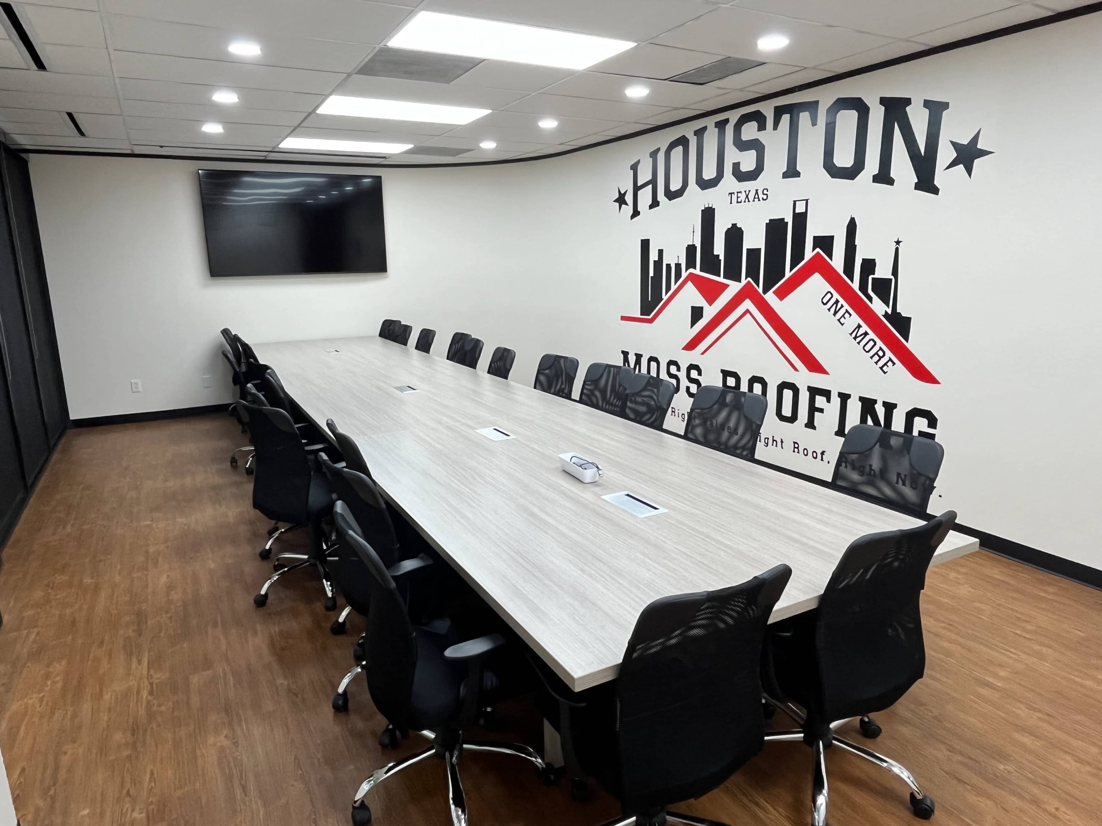 The image shows a large conference room with a long table surrounded by black chairs, featuring a wall with a large mural that includes the words "HOUSTON TEXAS" and "MOSS ROOFING."