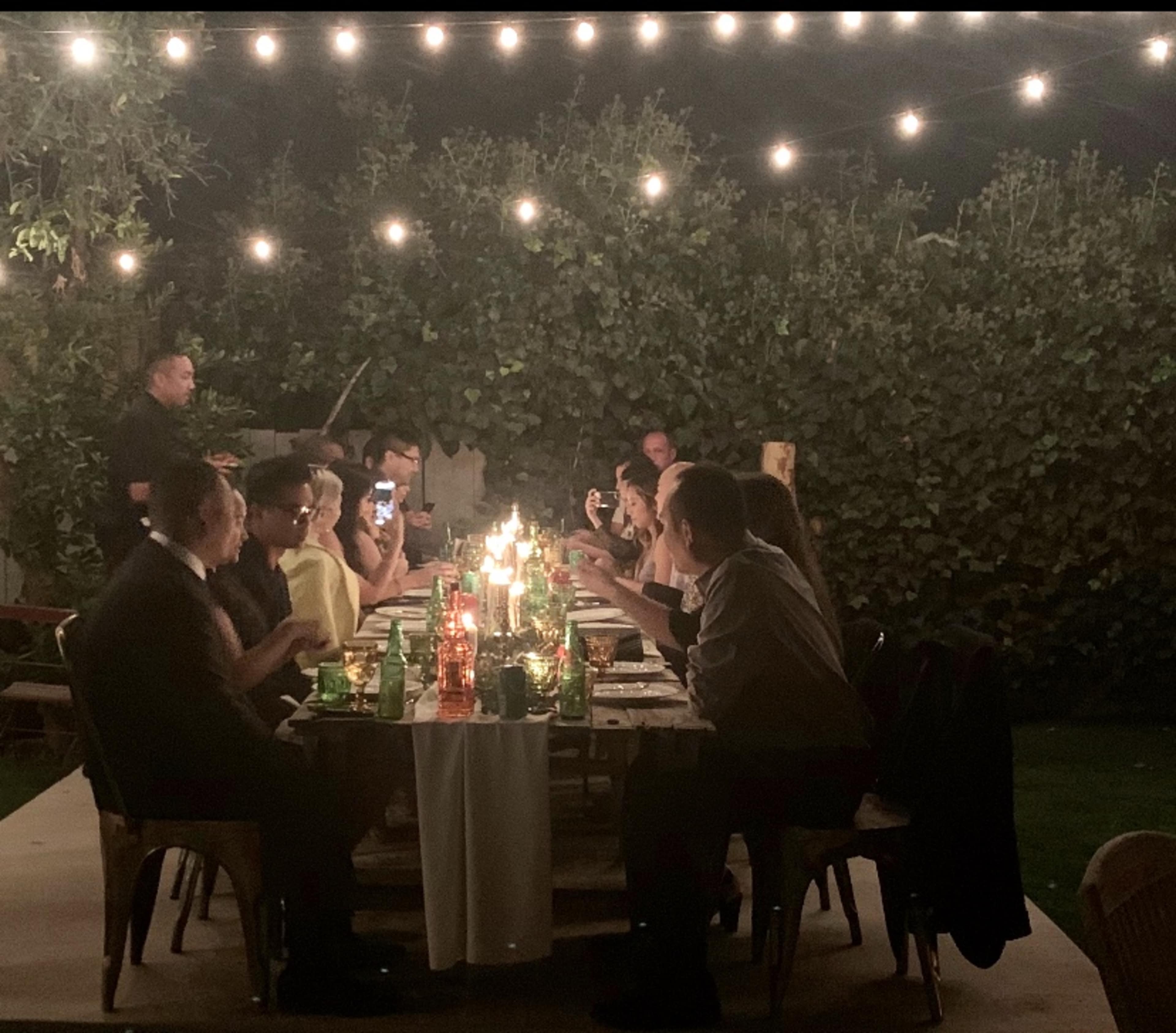 A group of people are seated at a long table outdoors, illuminated by string lights, while enjoying a meal and engaging in conversation.