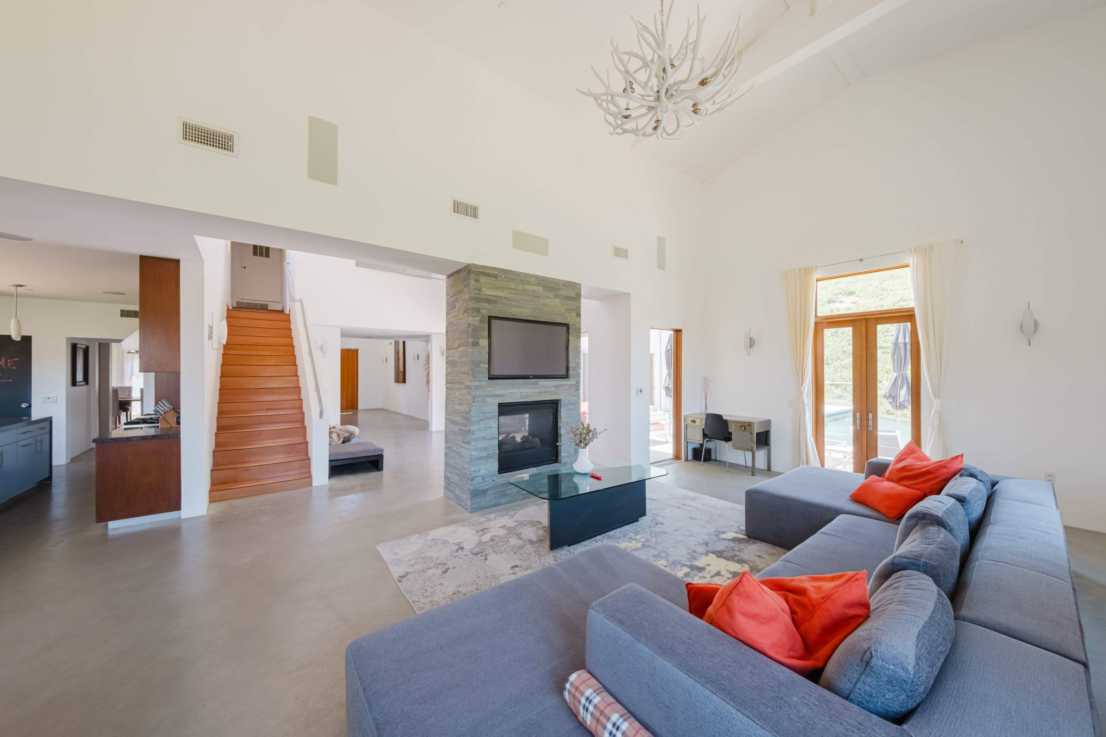 A spacious living room with a modern design, featuring a sectional sofa, a glass coffee table, a stone fireplace, and large windows that provide natural light.