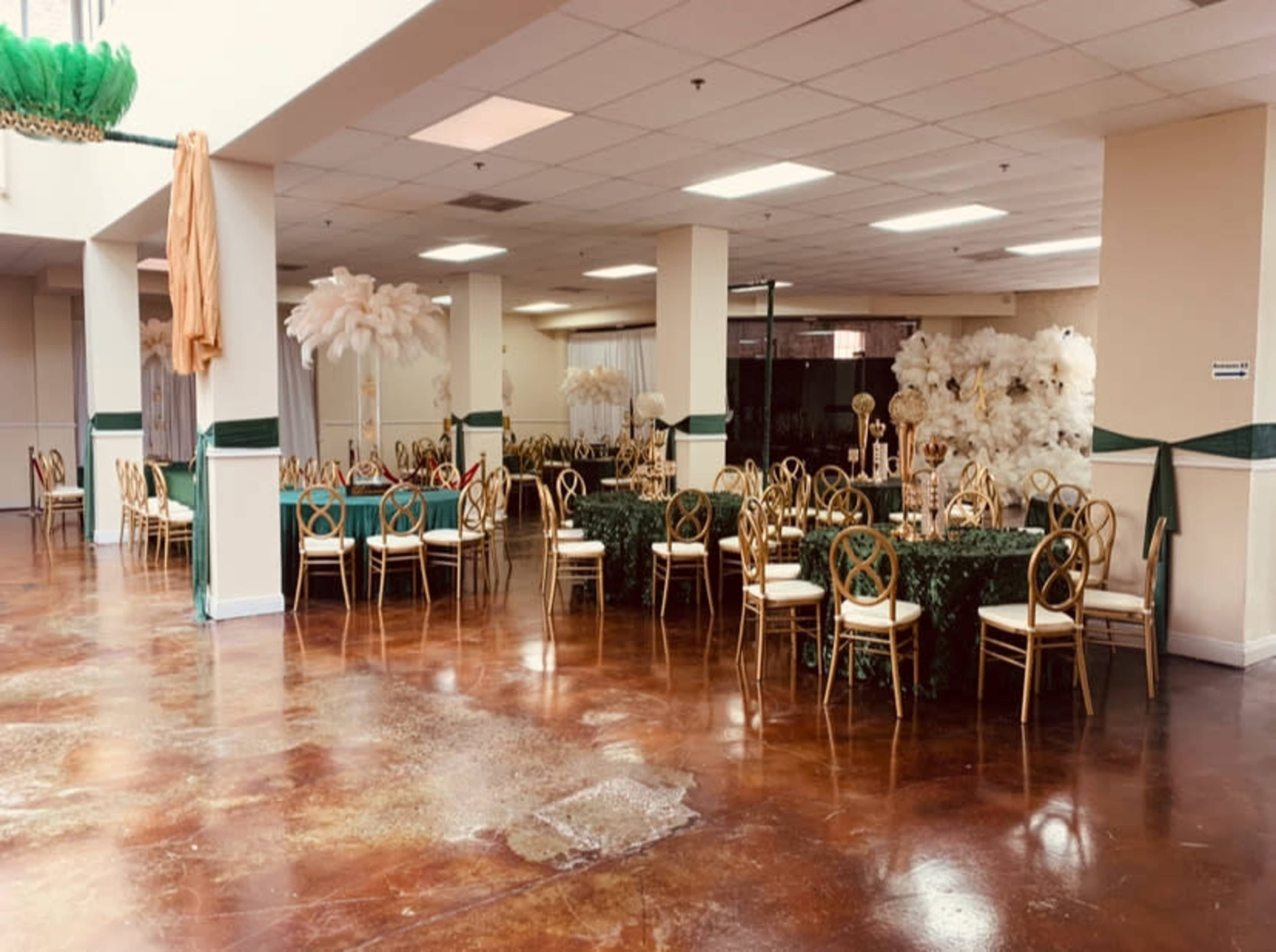 The image shows a banquet hall with round tables covered in green tablecloths and decorated with large white floral centerpieces, surrounded by gold chairs.