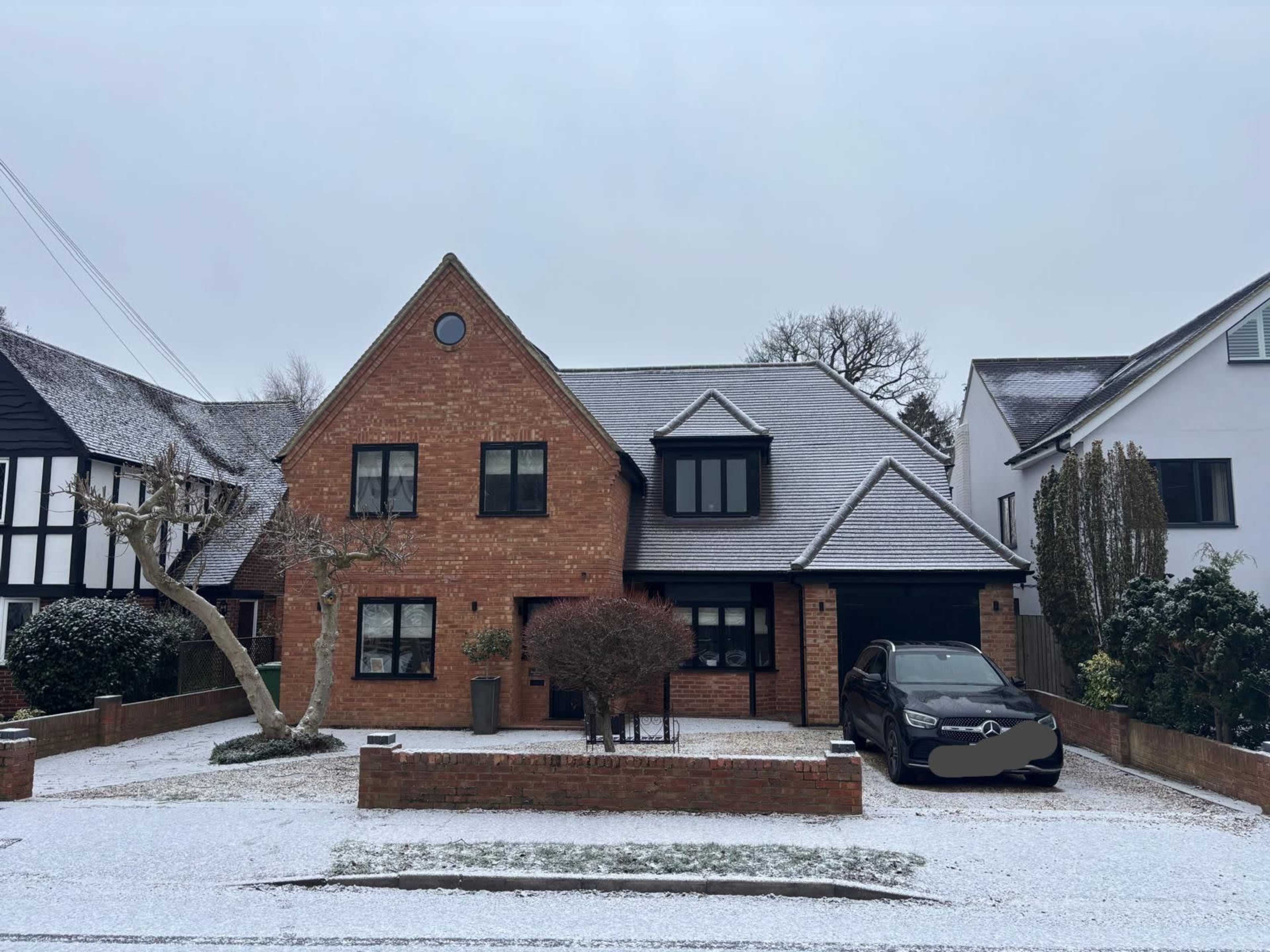 A brick house with a black roof and a car parked in the driveway is covered in a layer of snow.