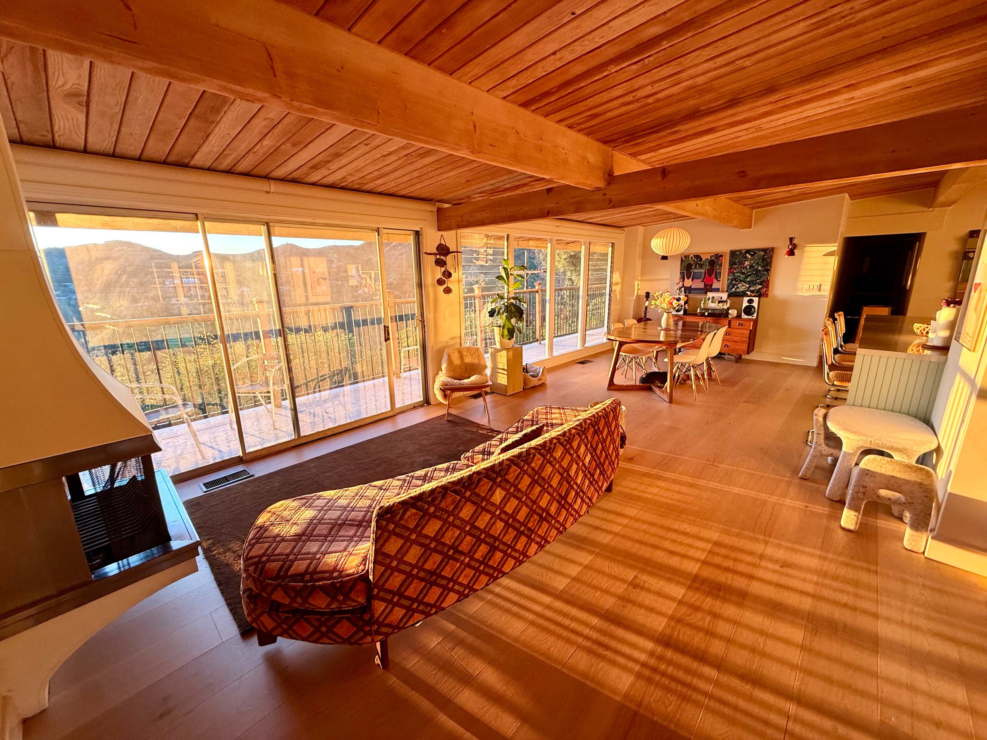 The interior of a modern living room features wooden beams, large windows with a view of mountains, and a mix of stylish furniture.