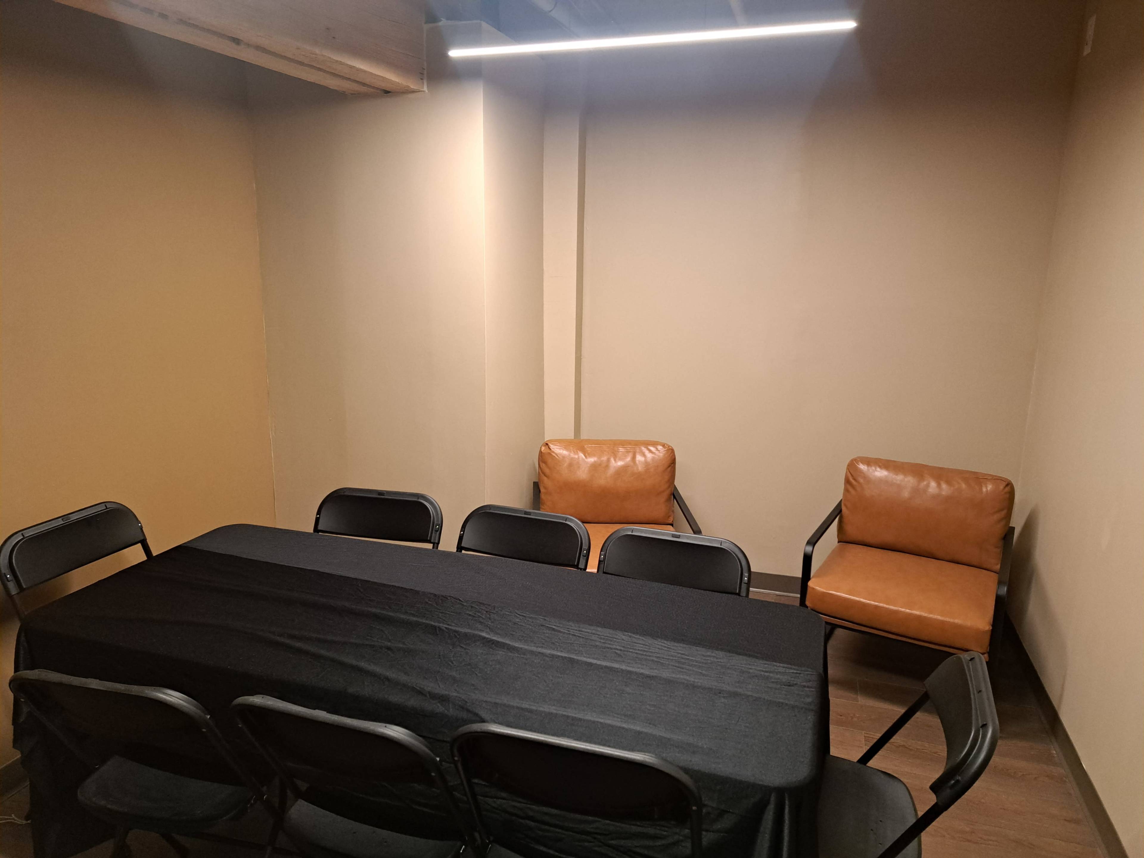 The image depicts a small, minimalistic meeting room with a black table, several black folding chairs, and two brown upholstered chairs against the wall.