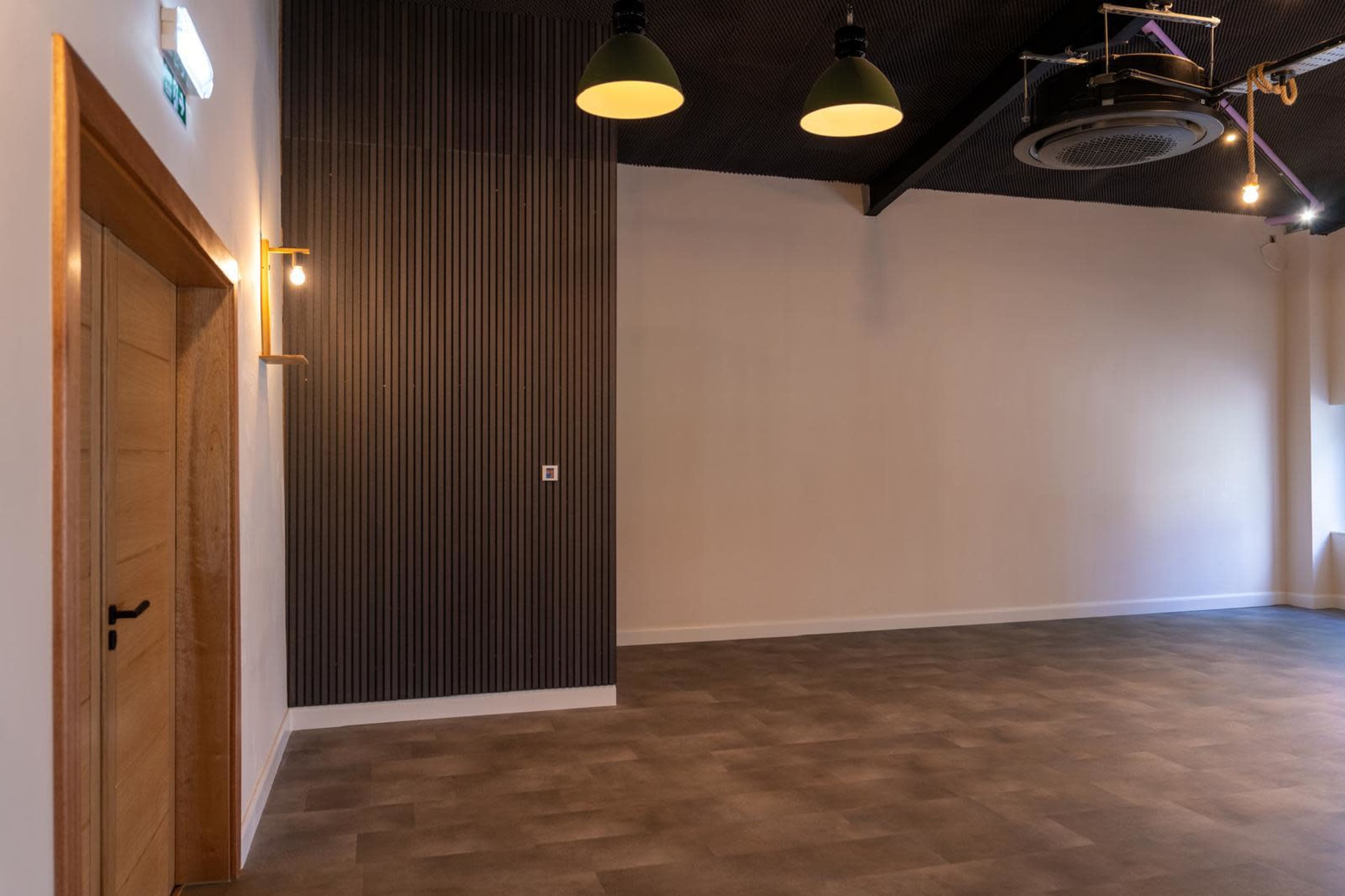The image shows a spacious, empty room with light-colored walls, a wooden door, and industrial-style hanging lights.