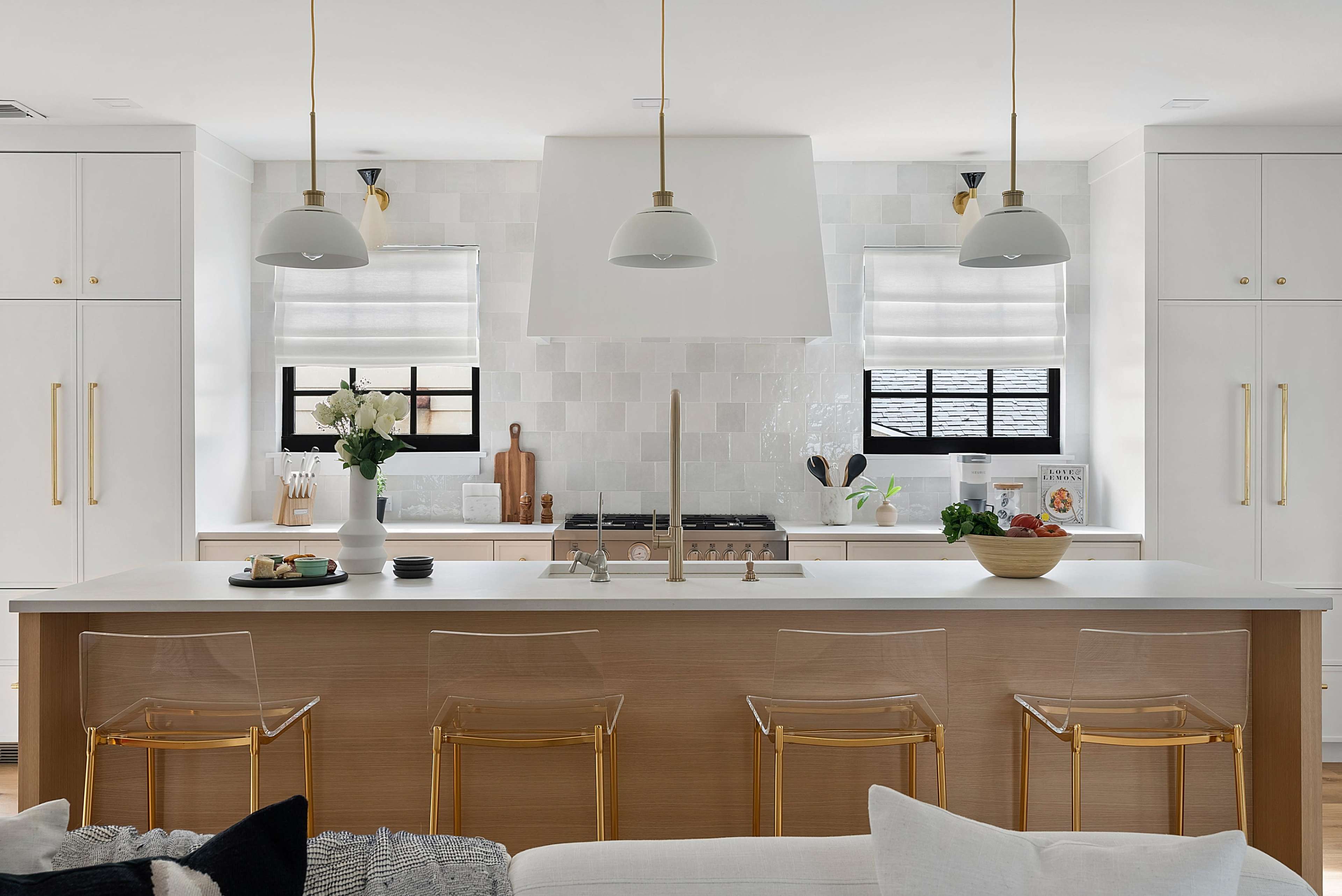 The kitchen features a large island with transparent stools, white cabinets, and a tiled backsplash, illuminated by pendant lights.
