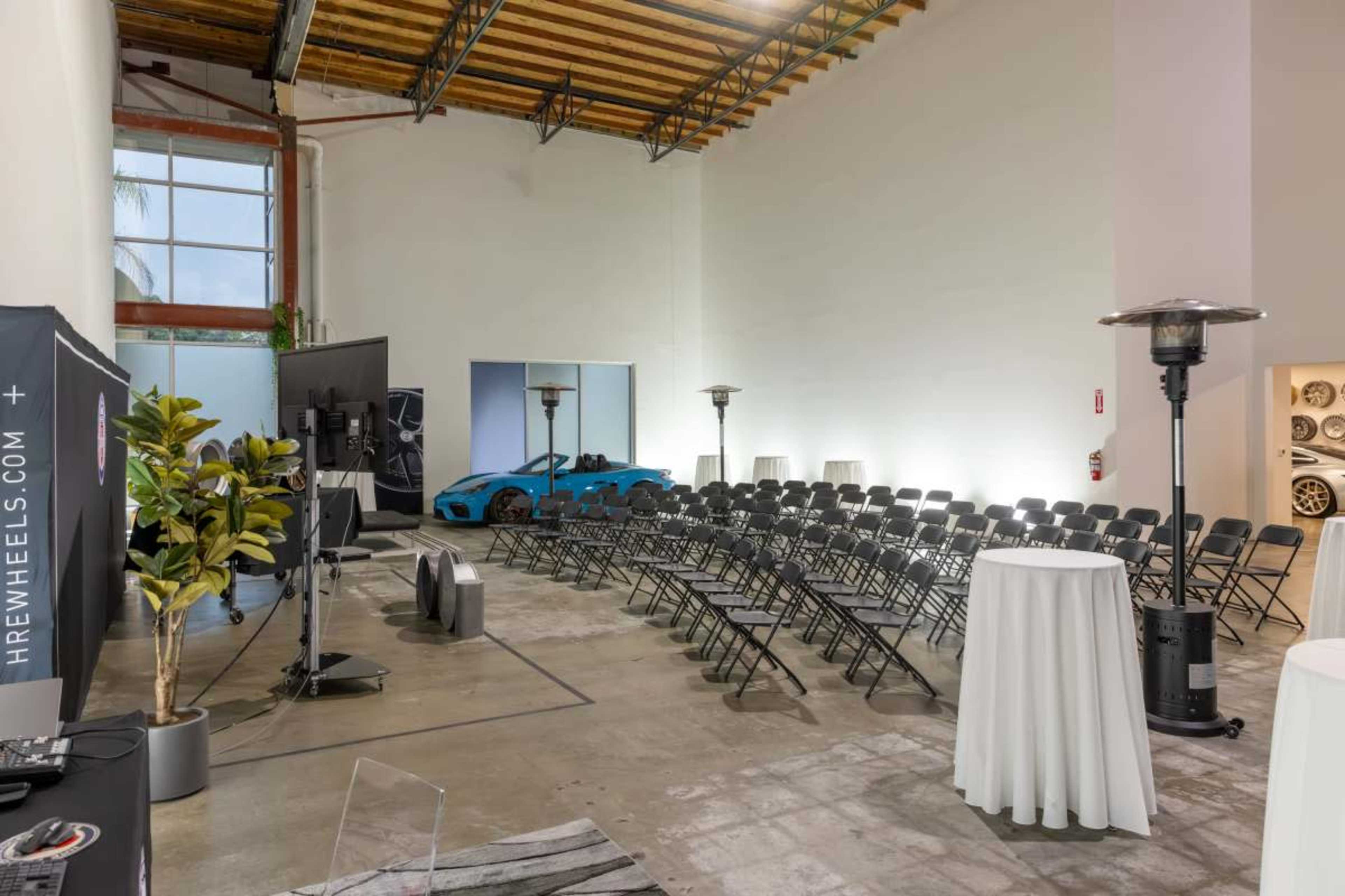 The image shows a large, industrial-style room set up for an event, featuring rows of black chairs, two tall heaters, and a blue sports car parked towards the back.
