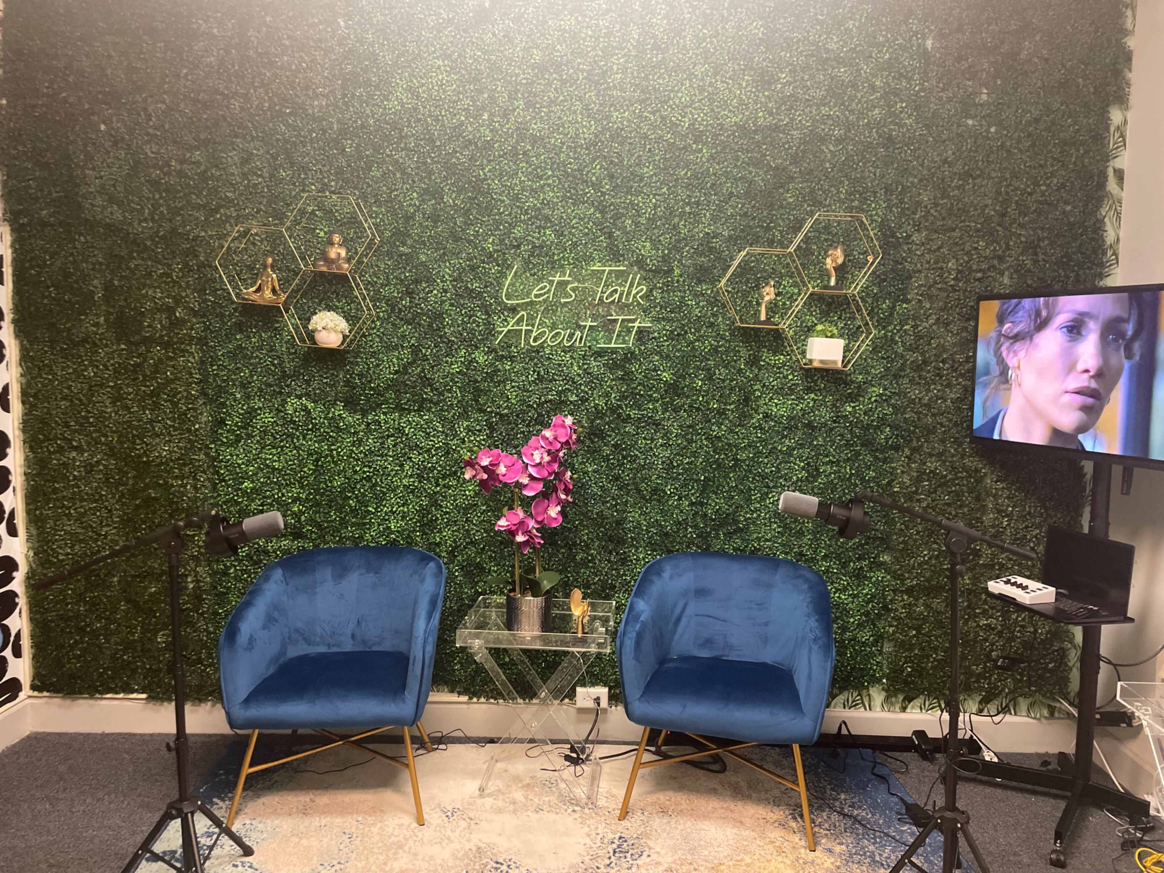 A small studio setup with two blue chairs facing each other, a glass table with a pink flower arrangement, and a green hedge wall featuring decorative hexagonal shelves.