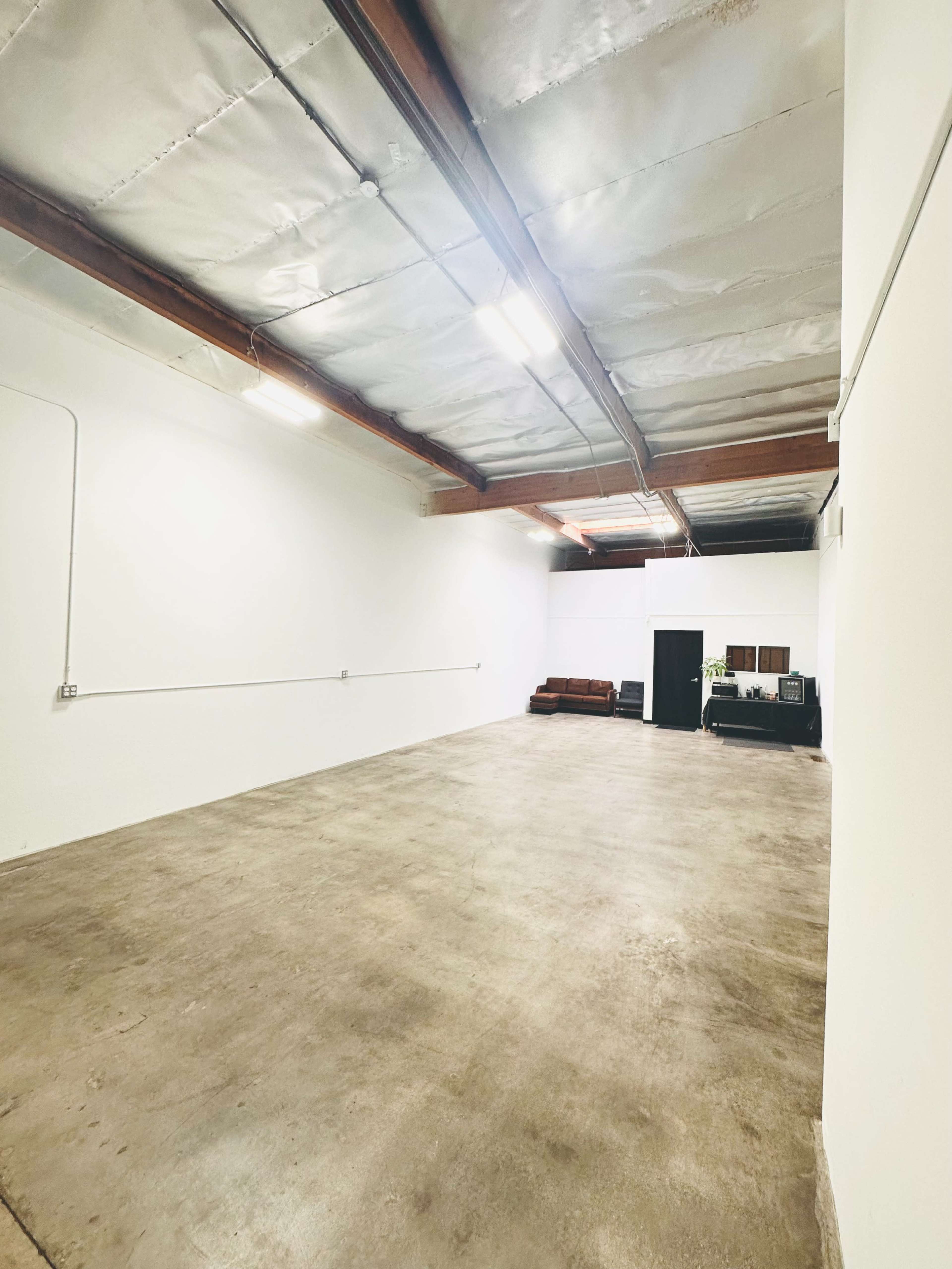 A large, empty room with concrete flooring and white walls, featuring a few pieces of furniture along one side.