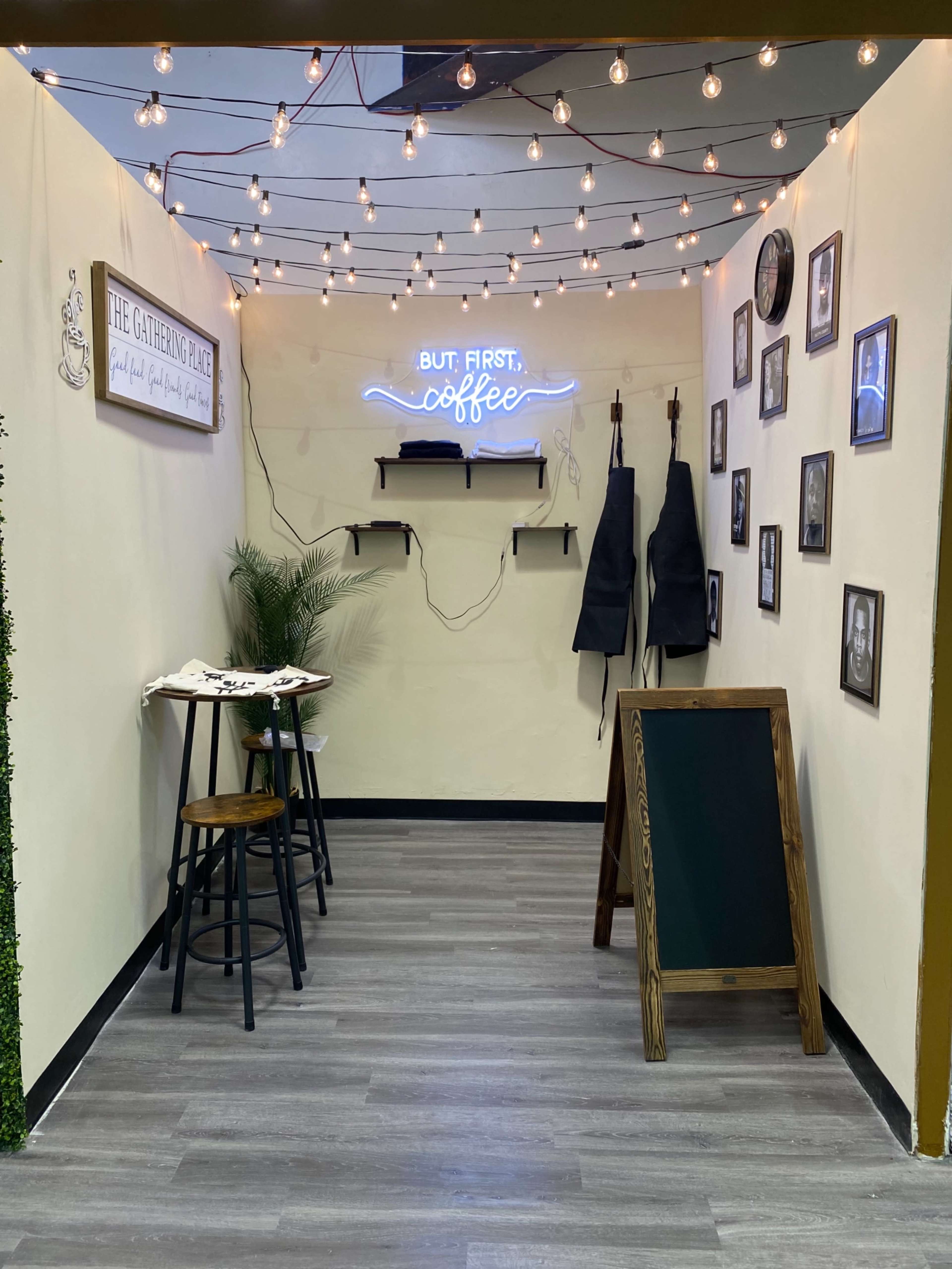 A small, cozy coffee corner with string lights, a neon sign that says "BUT FIRST, coffee," two high stools, and a wall decorated with framed photos.