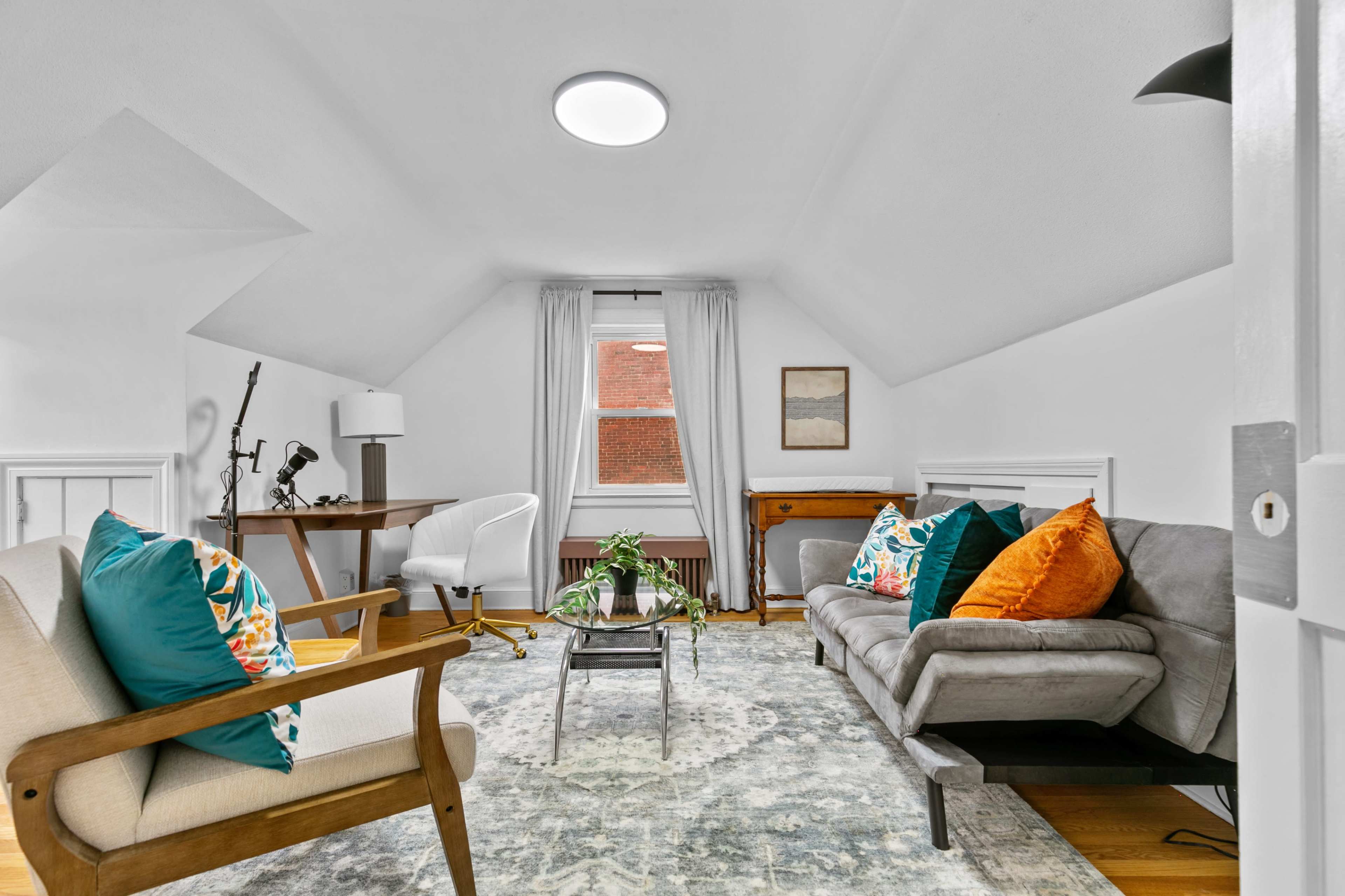 A cozy living space with a sloped ceiling, featuring two armchairs and a sofa arranged around a central coffee table, a desk in one corner, and a window with light curtains.