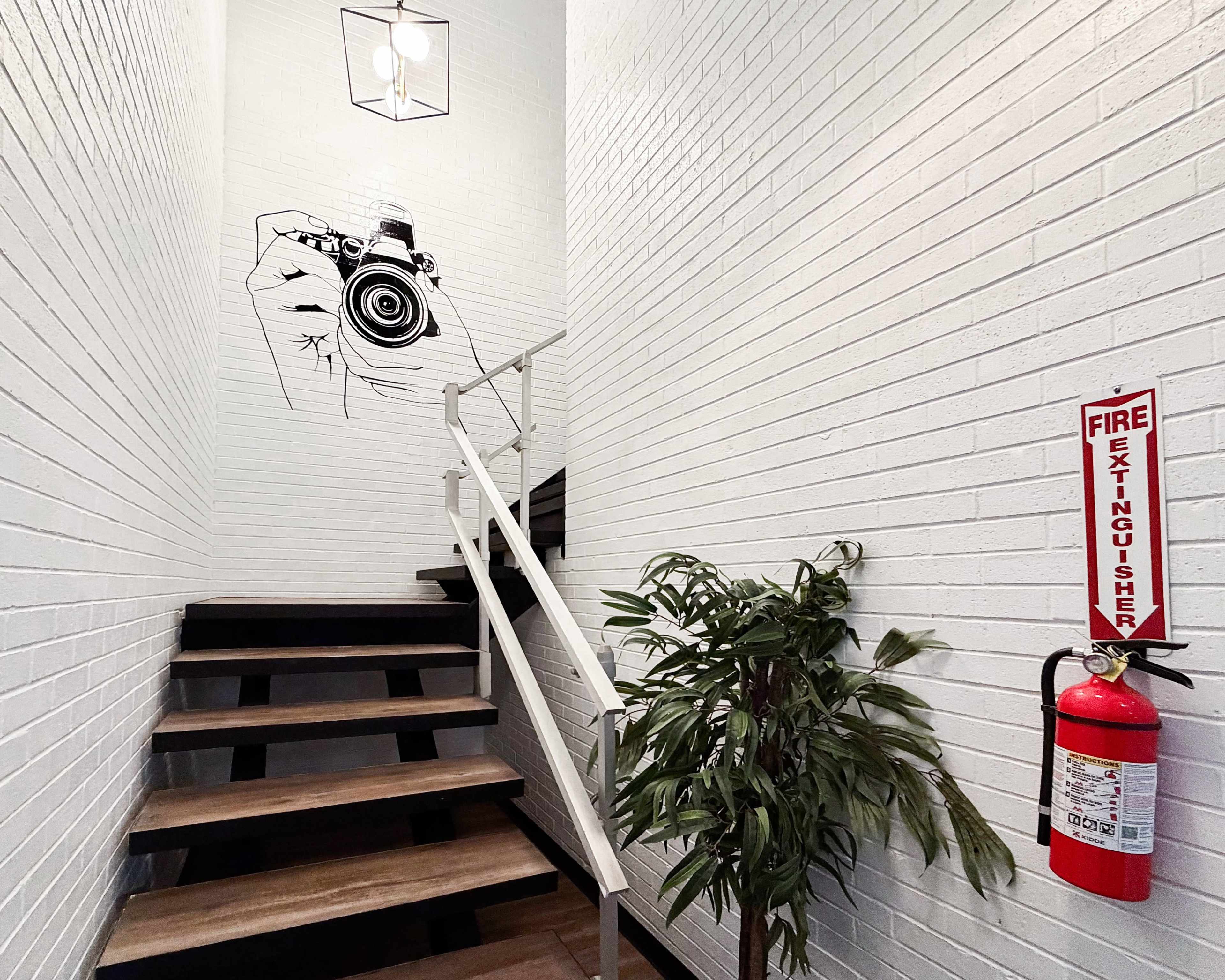 A staircase with wooden steps leads up to a landing adorned with a camera mural on the white brick wall, accompanied by a fire extinguisher and a potted plant.