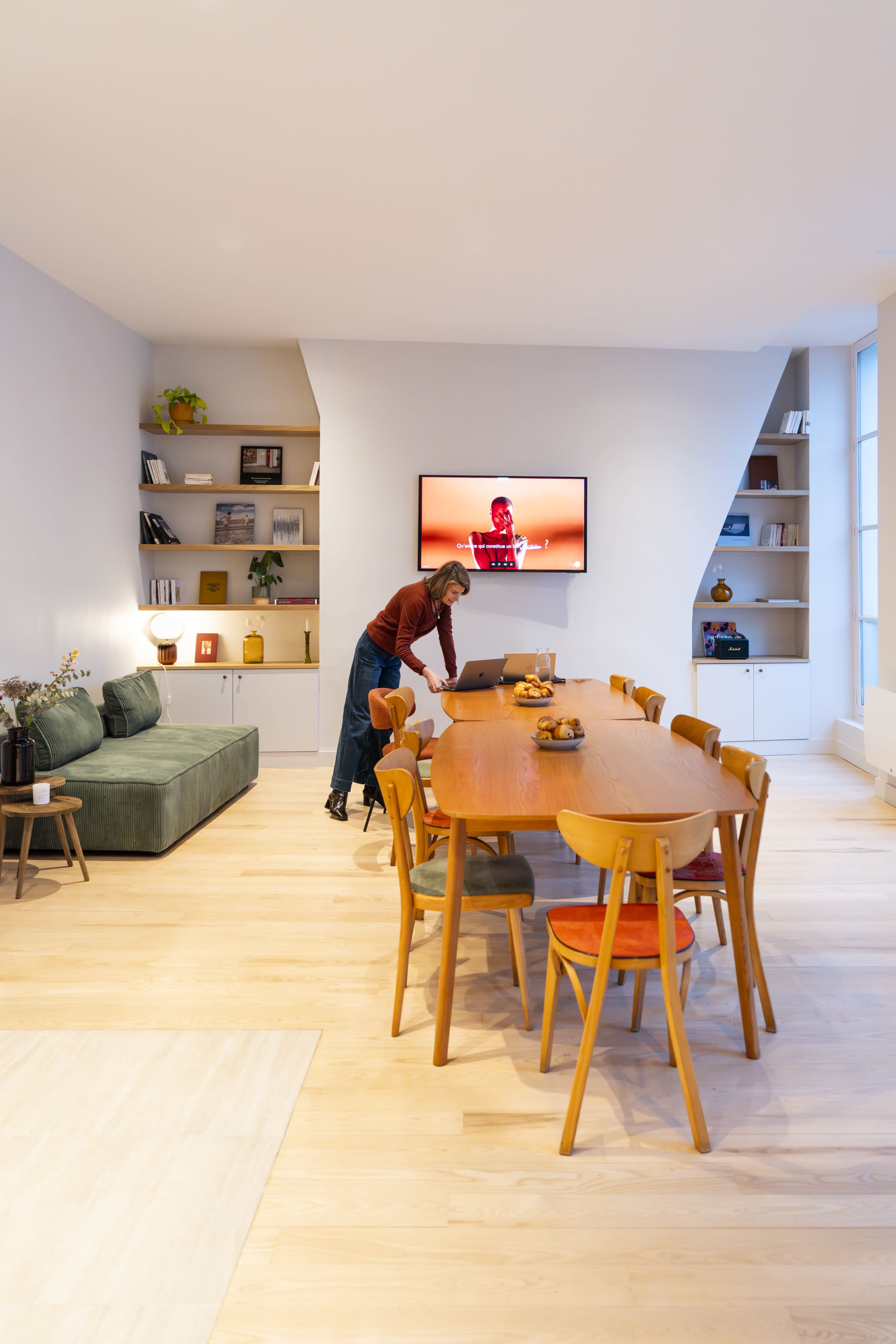 A woman is setting the wooden dining table in a modern living space with a green sofa and a TV on the wall.