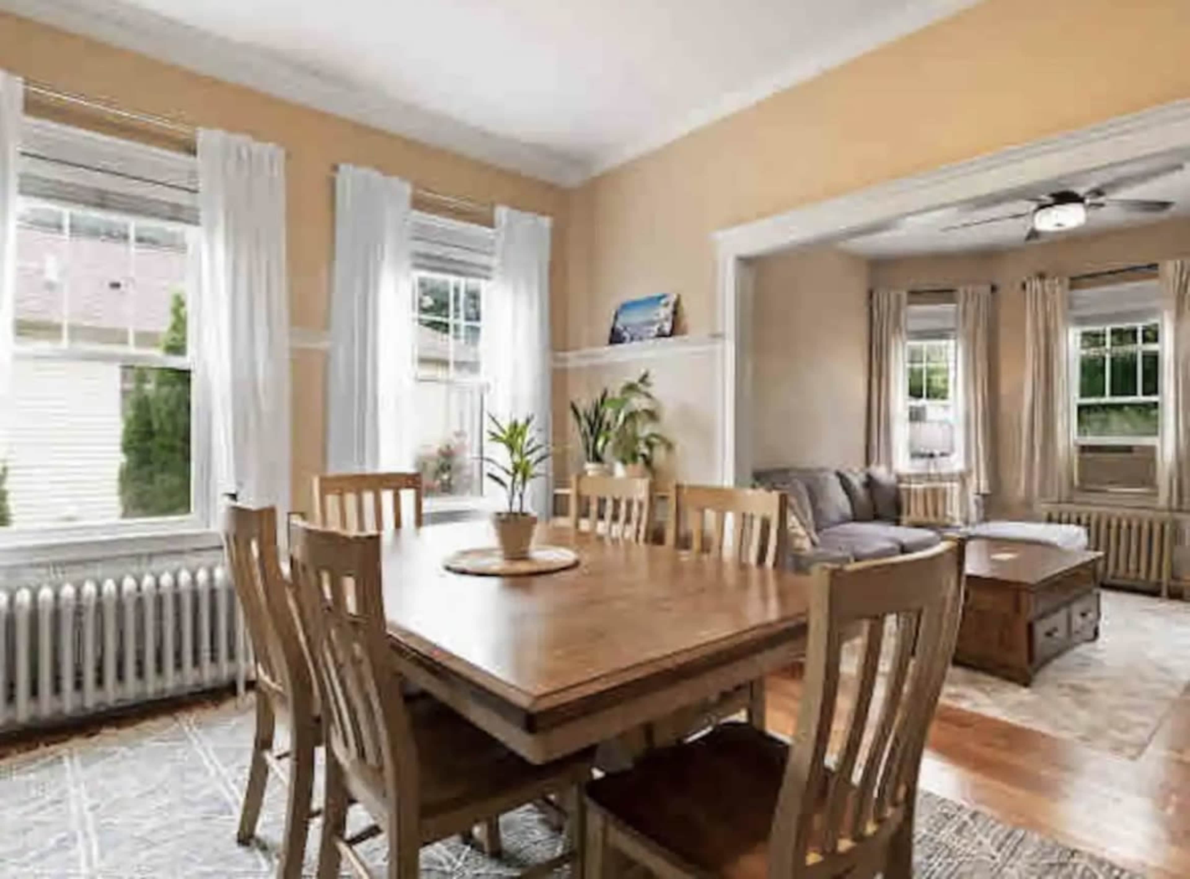 A dining area with a wooden table and chairs, large windows allowing natural light, and a separate living space with a couch and coffee table in the background.