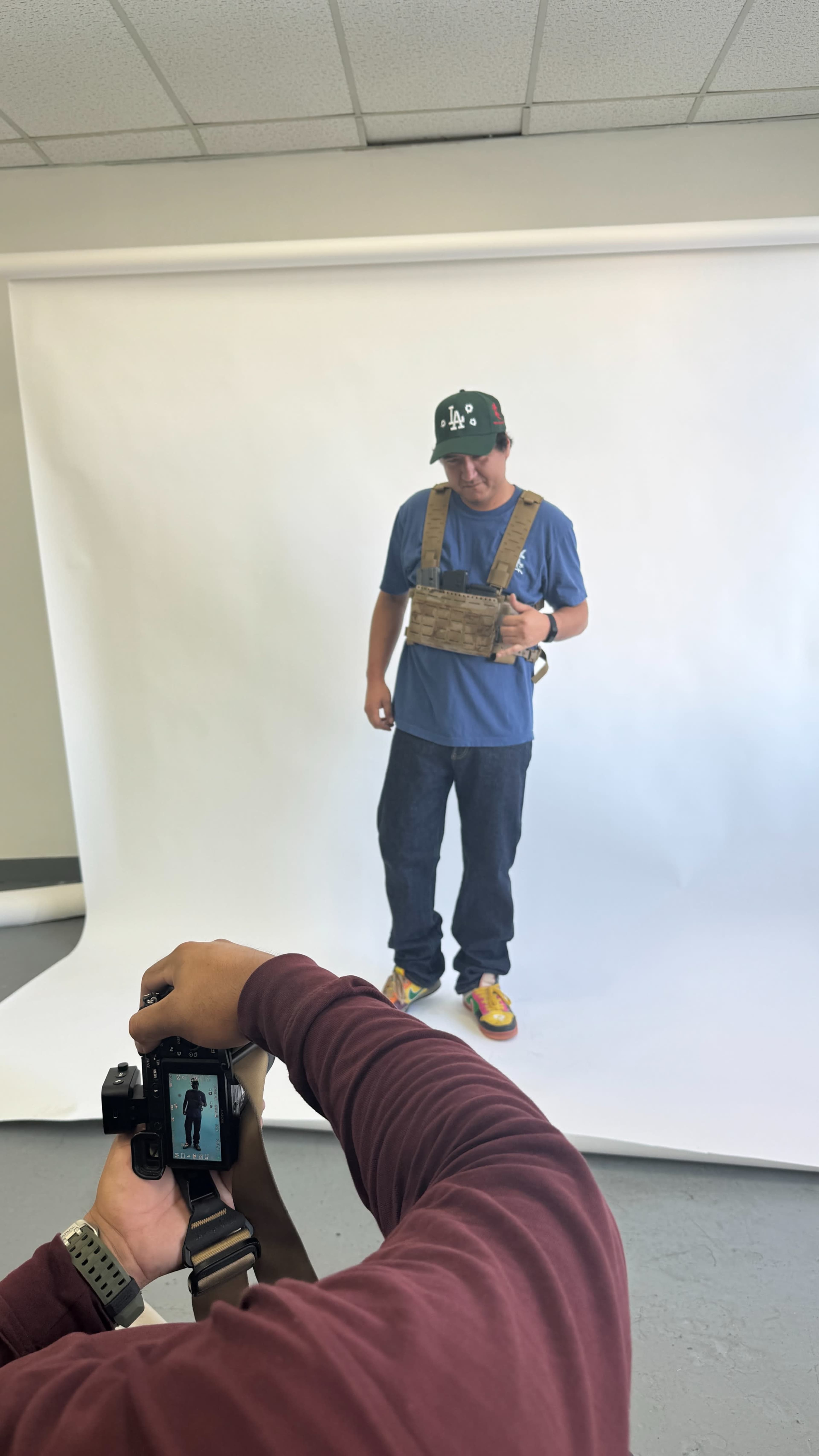 A person wearing a tactical vest and casual clothing poses for a photo in front of a white backdrop while another person holds a camera.