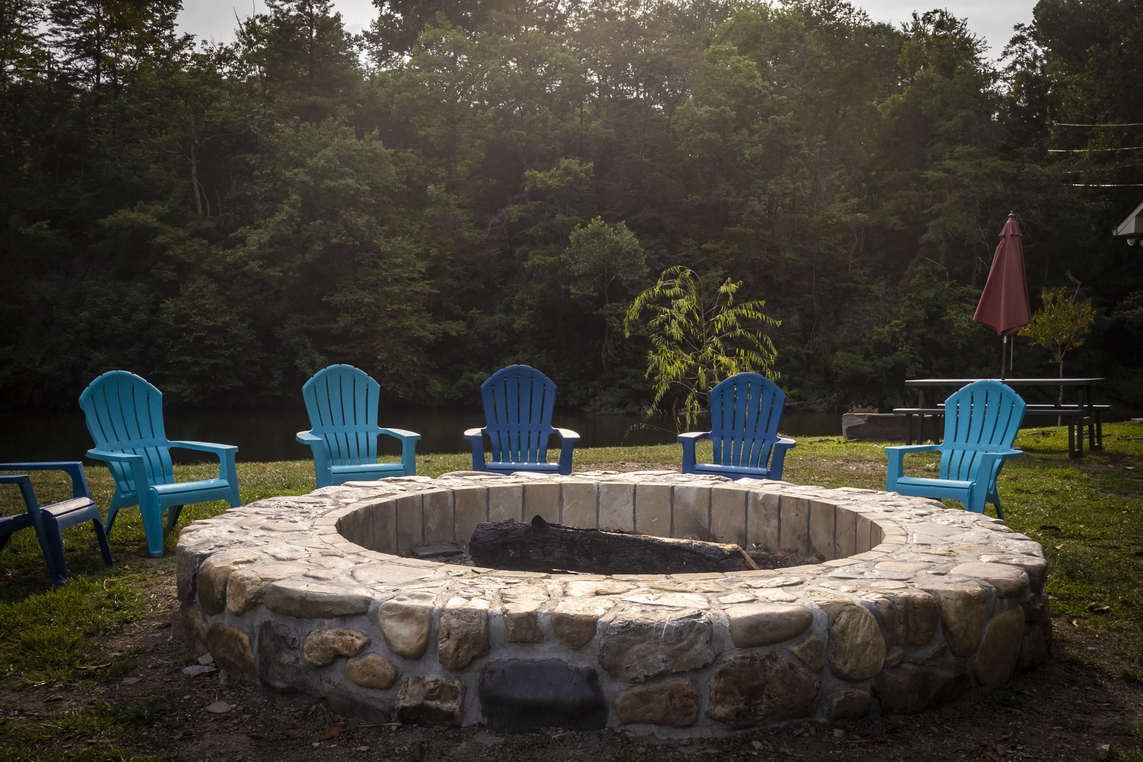 A circular stone fire pit is surrounded by five blue adirondack chairs in a wooded area near a body of water.