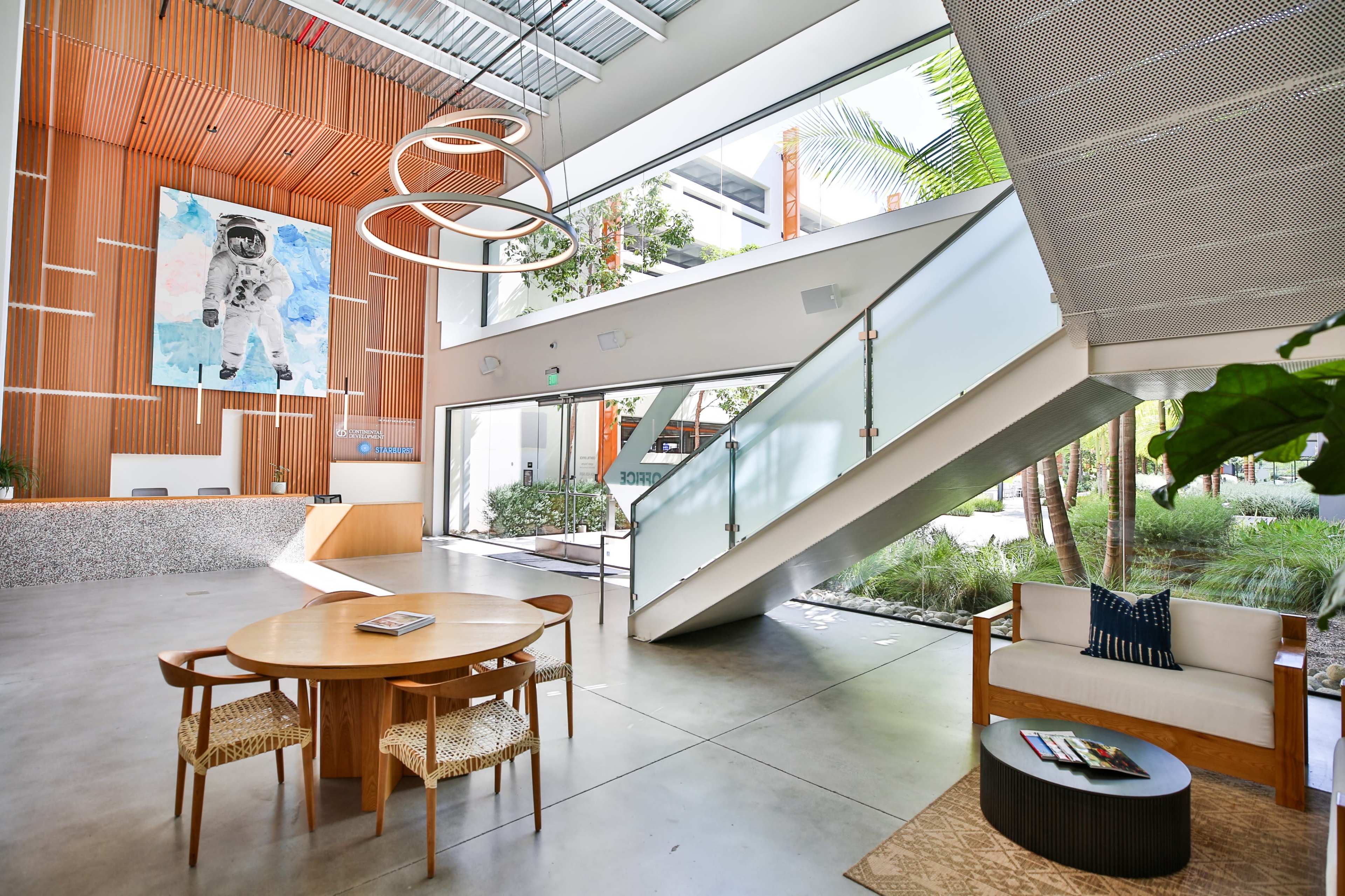 A spacious lobby with a simple circular table, a couch, and a staircase leading to an upper level, characterized by large windows and a prominent artwork of an astronaut on the wall.