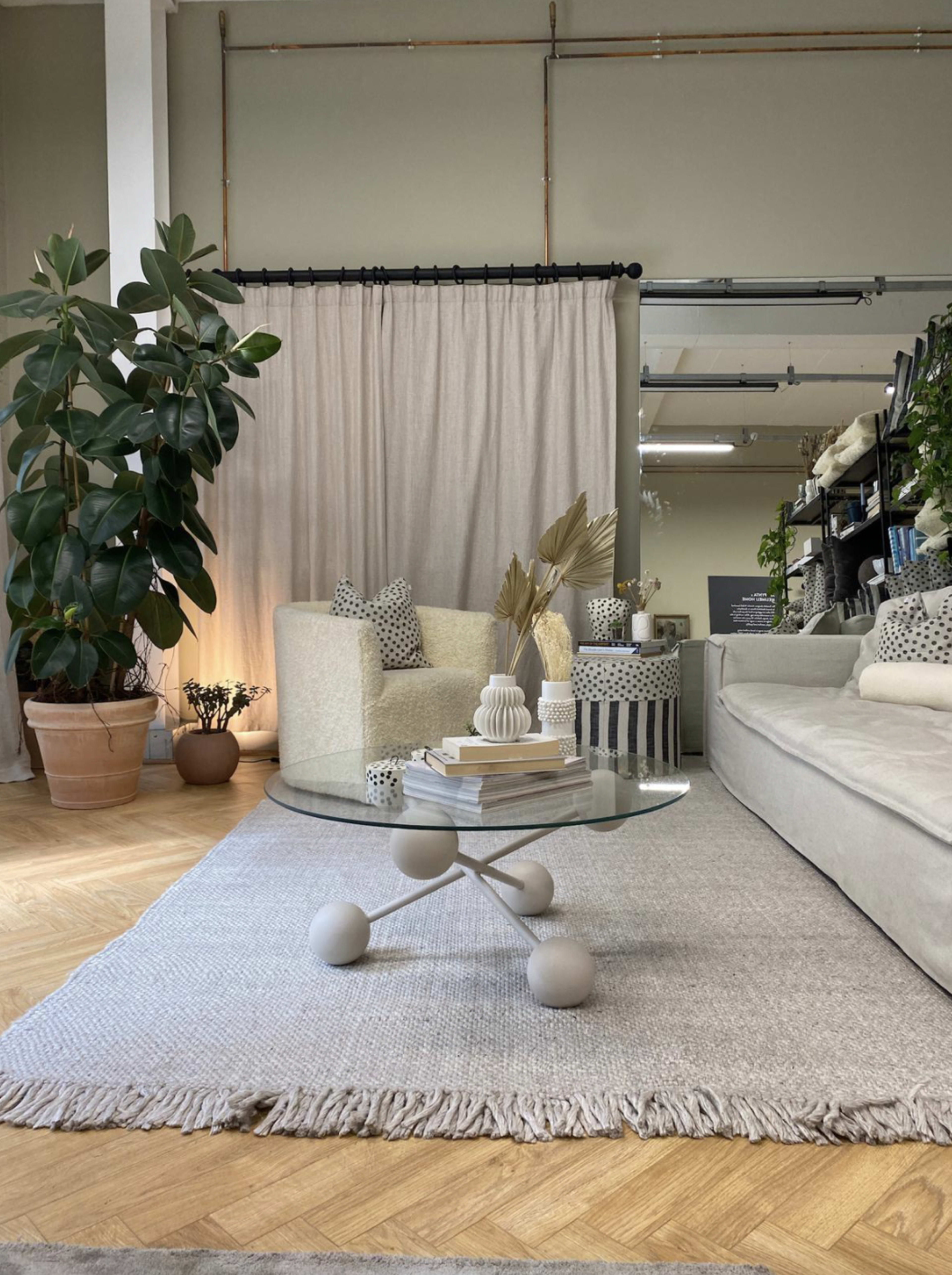 A cozy living space features a light-colored sofa, a glass coffee table with decorative items, and potted plants, set against a backdrop of soft curtains and shelving.