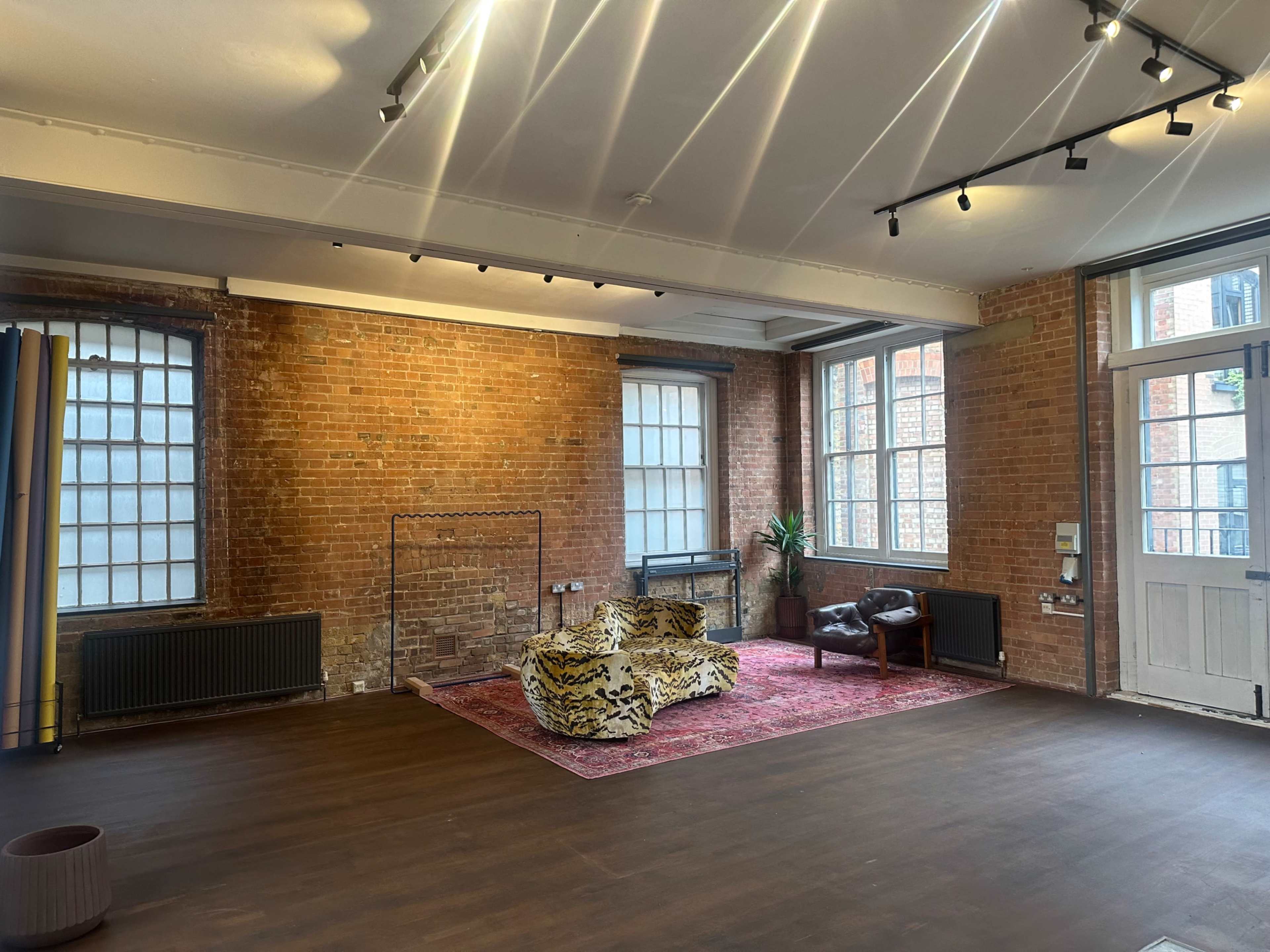 The image shows an empty room with exposed brick walls, large windows, a patterned sofa, and a rug in the center.