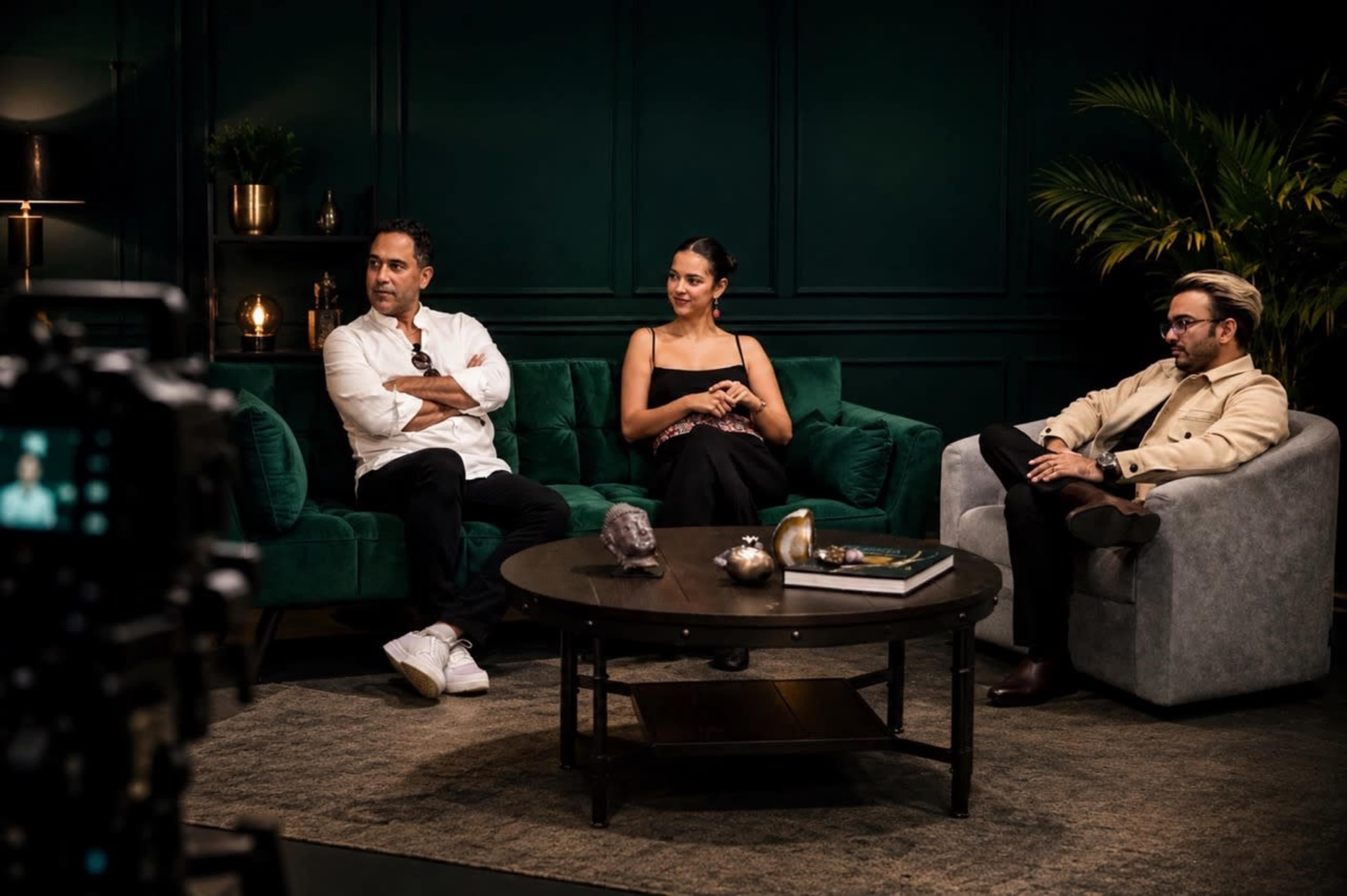 Three individuals sit on a green sofa in a stylish living room, engaged in conversation while a camera captures the scene.