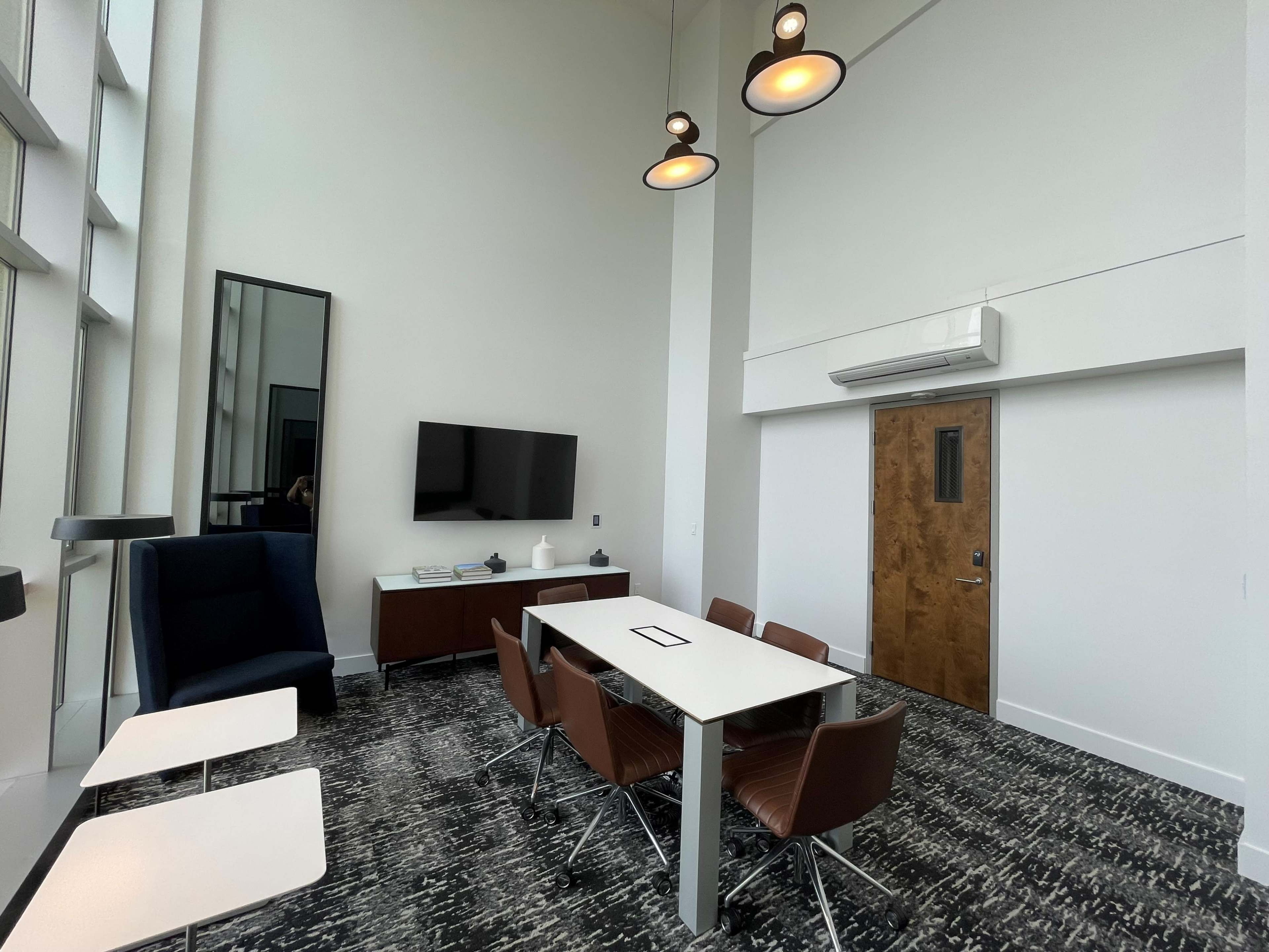 The image shows a modern conference room with a large table surrounded by chairs, a TV mounted on the wall, and large windows providing natural light.