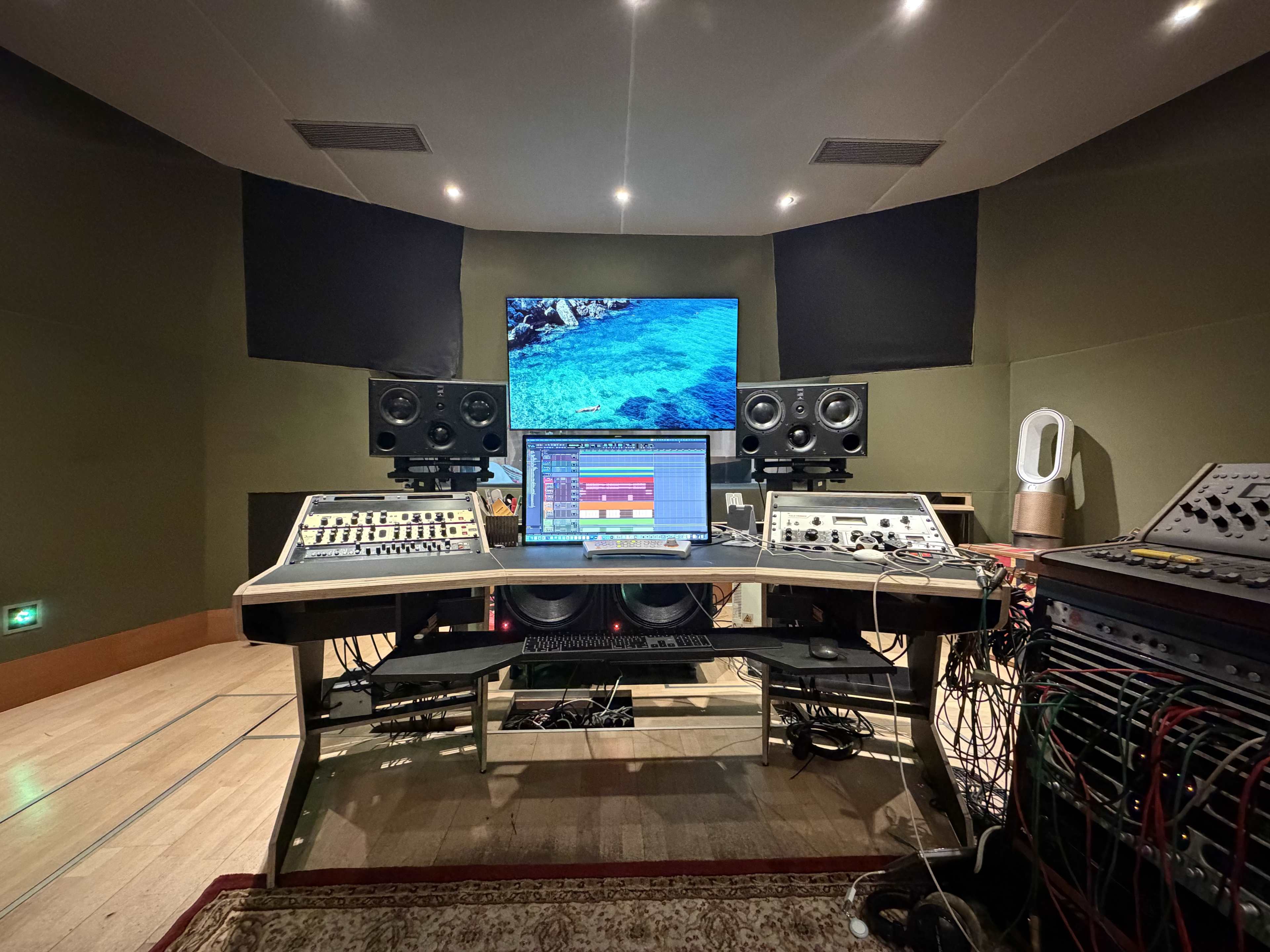 The image shows a music recording studio with various audio equipment, a computer display, and speakers arranged around a central mixing desk.