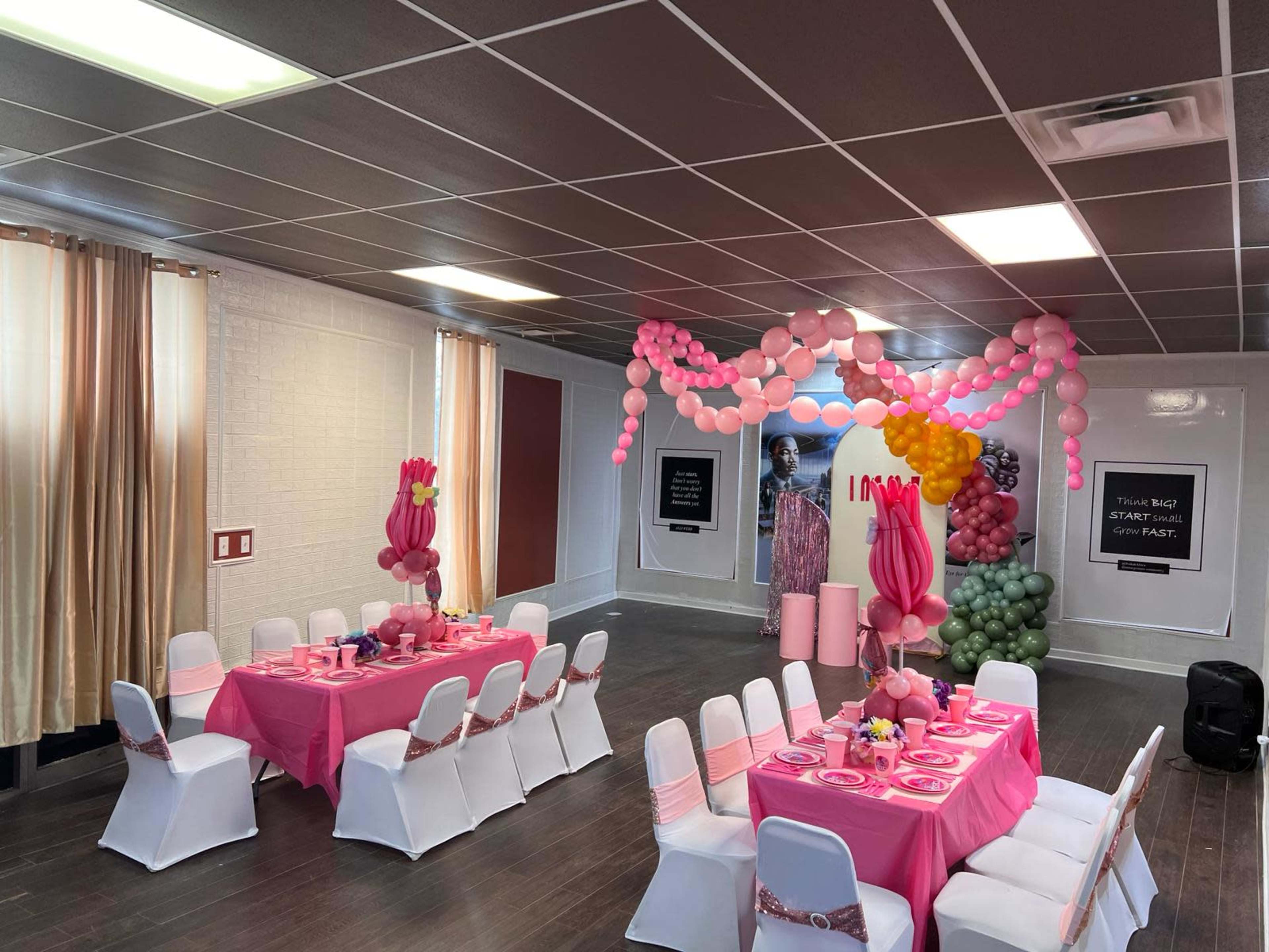 The image shows a decorated indoor party space featuring a table set with pink tableware and balloons arranged in a colorful backdrop.