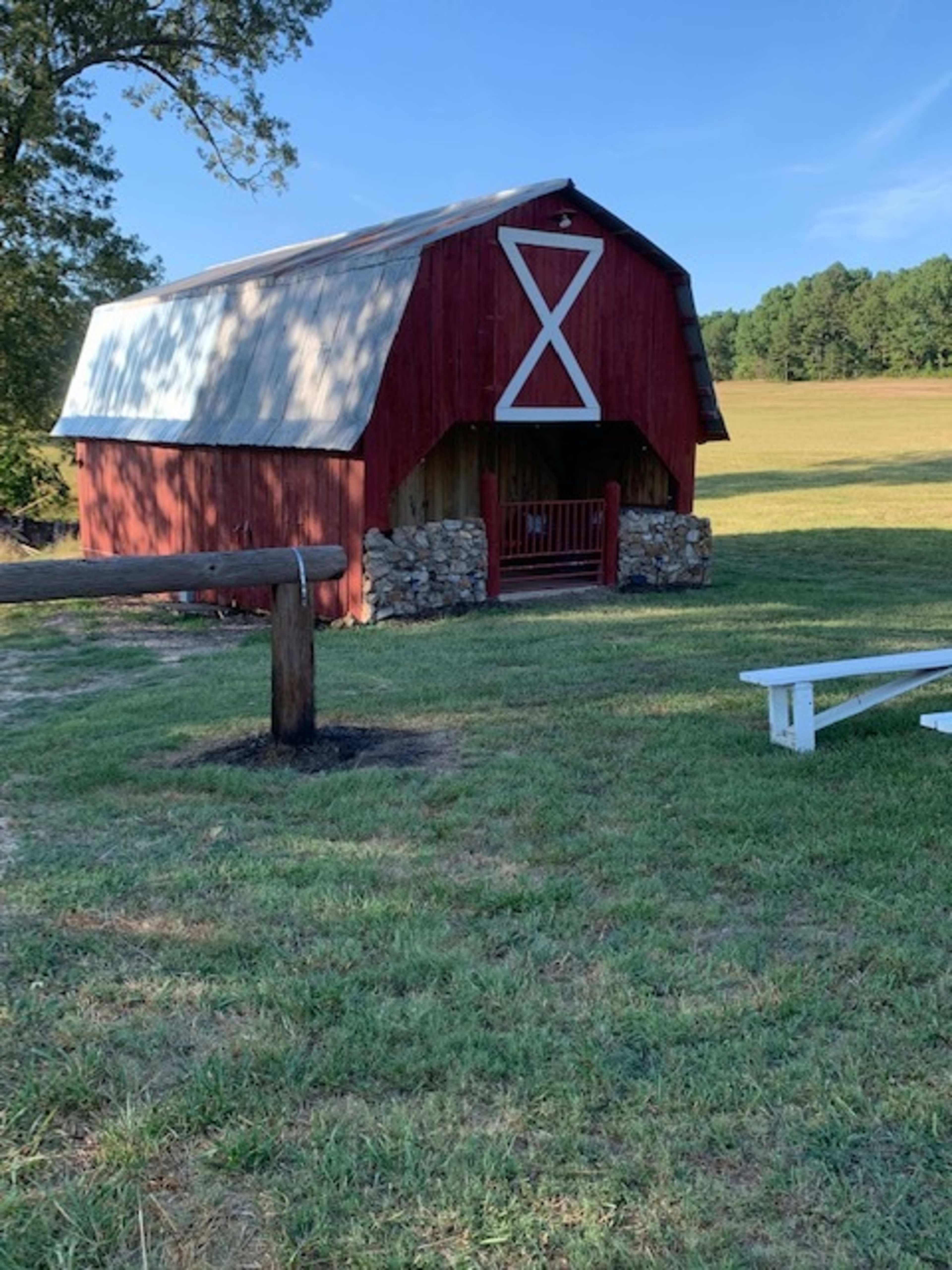 A red barn with a metal roof and a stone foundation is situated on a grassy field near a wooden fence.