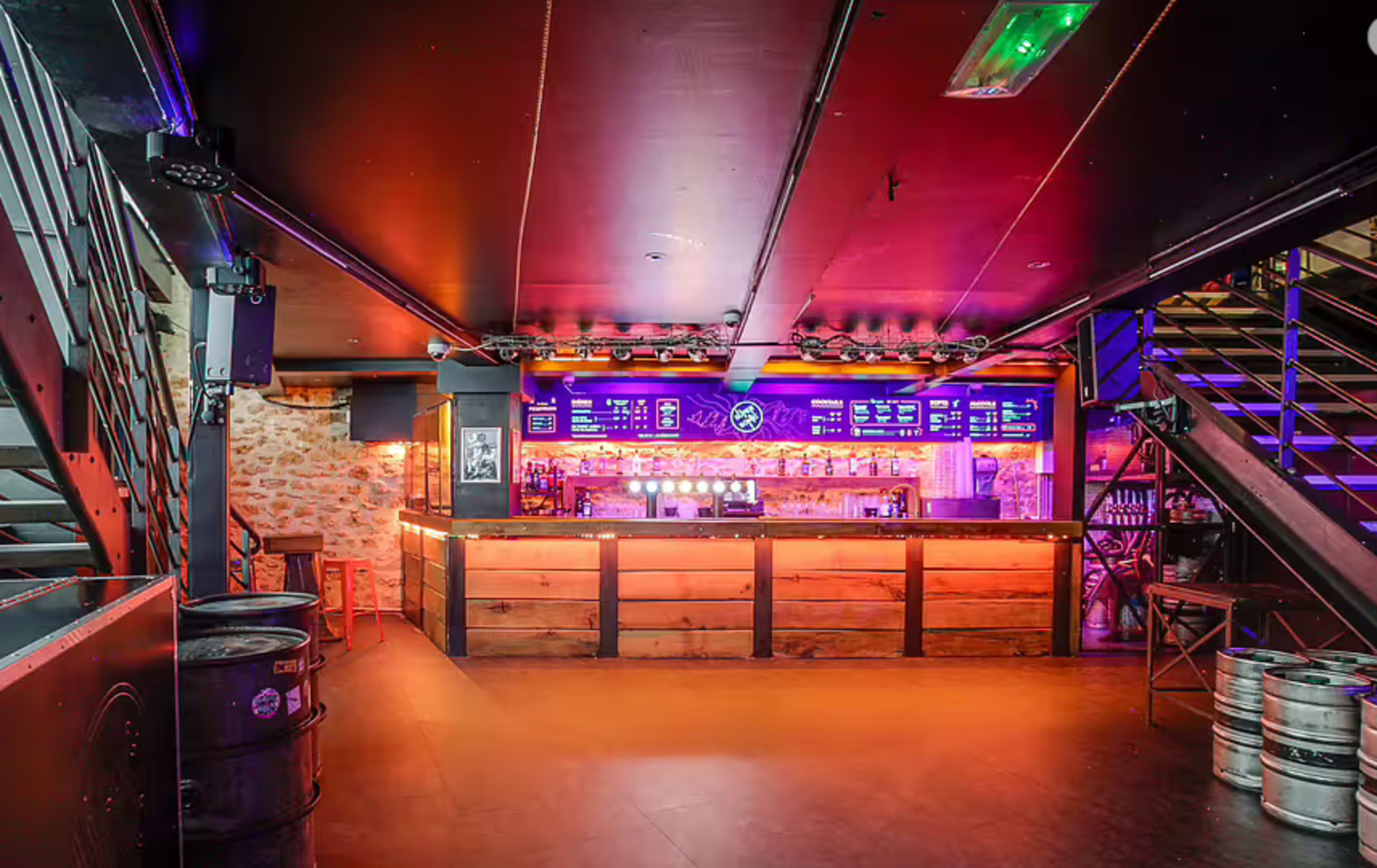 The image shows a modern bar interior with purple and orange lighting, a wooden bar counter, and metal staircase access.