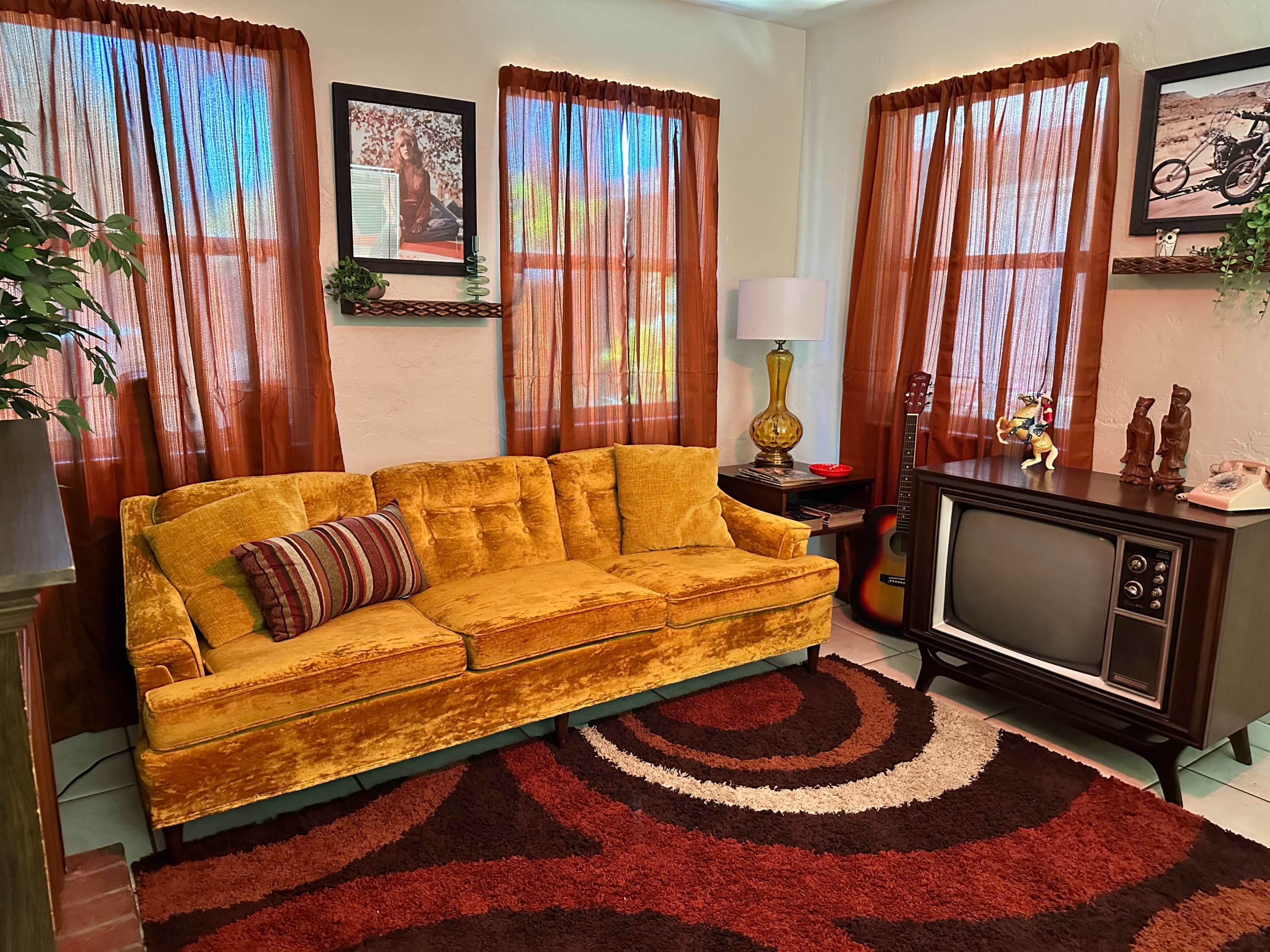 The room features a yellow velvet couch, vintage television set, and decorative elements, all arranged with brown curtains and a circular rug.