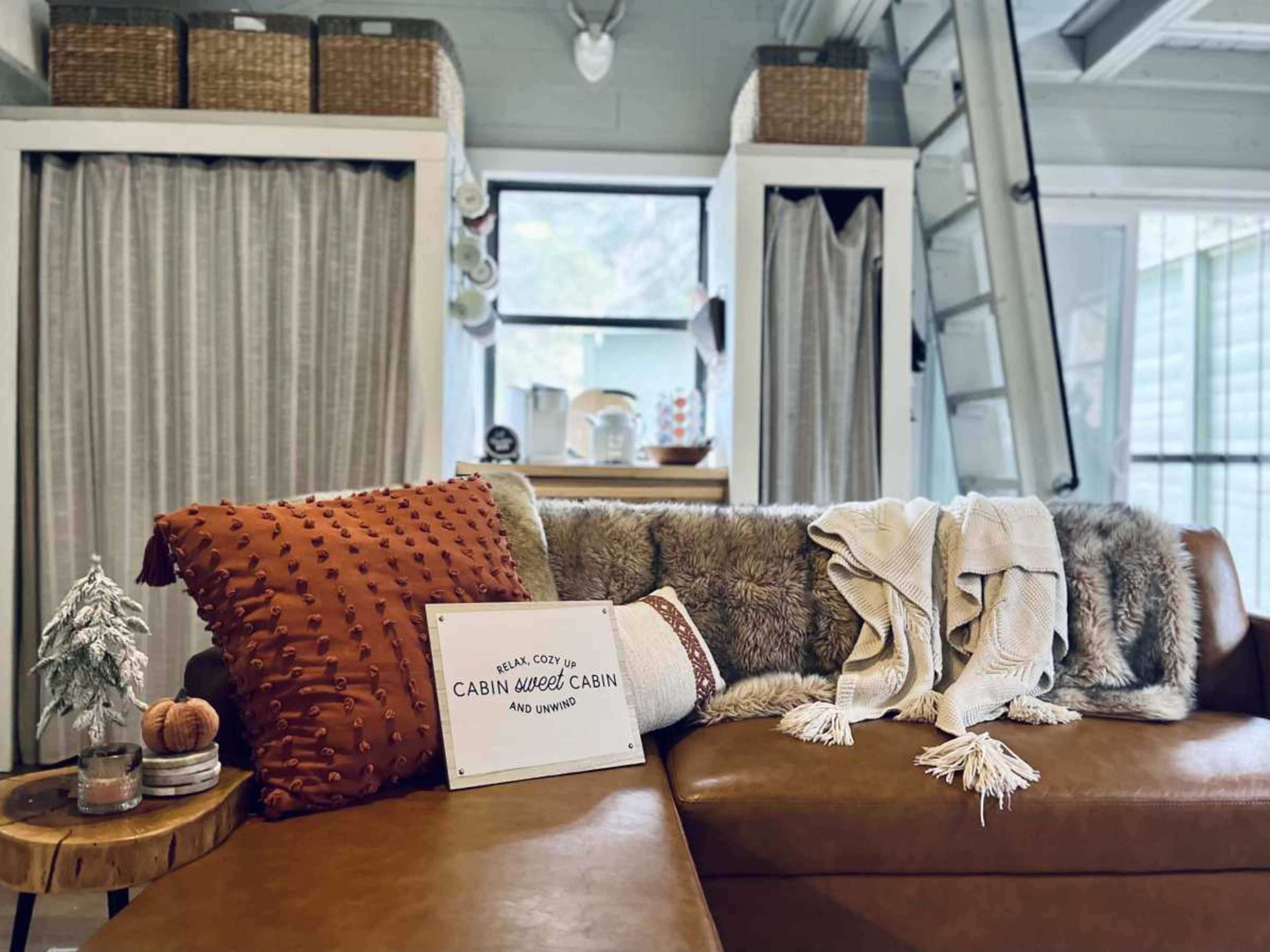 A cozy living room with a leather sofa adorned with decorative pillows and a throw blanket, alongside a sign that reads "CABIN WEED CABIN."