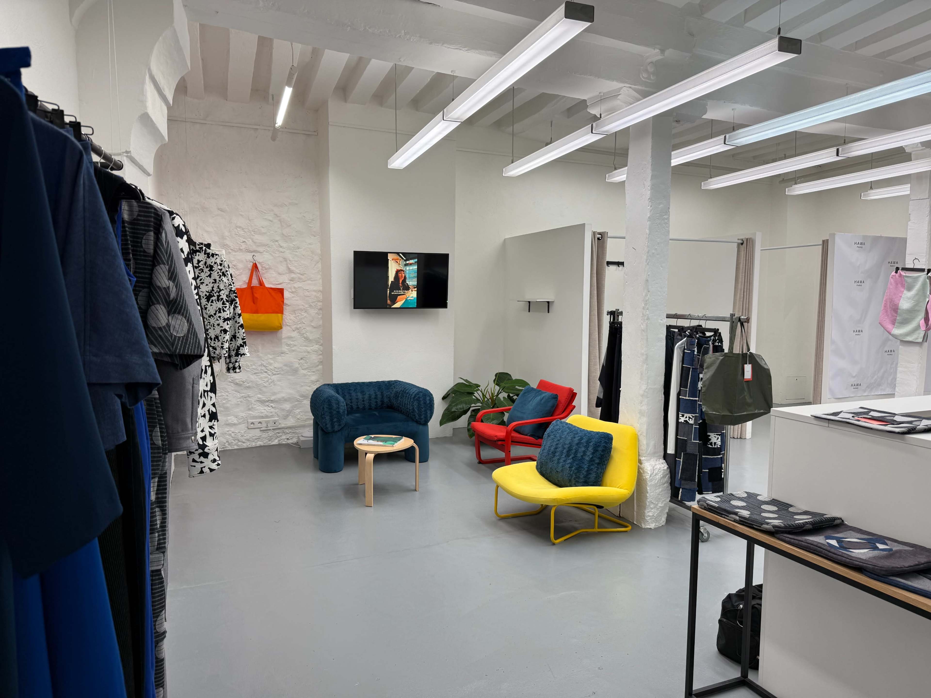 The image depicts a modern clothing store interior featuring colorful seating areas, hanging garments on racks, and a television mounted on the wall.