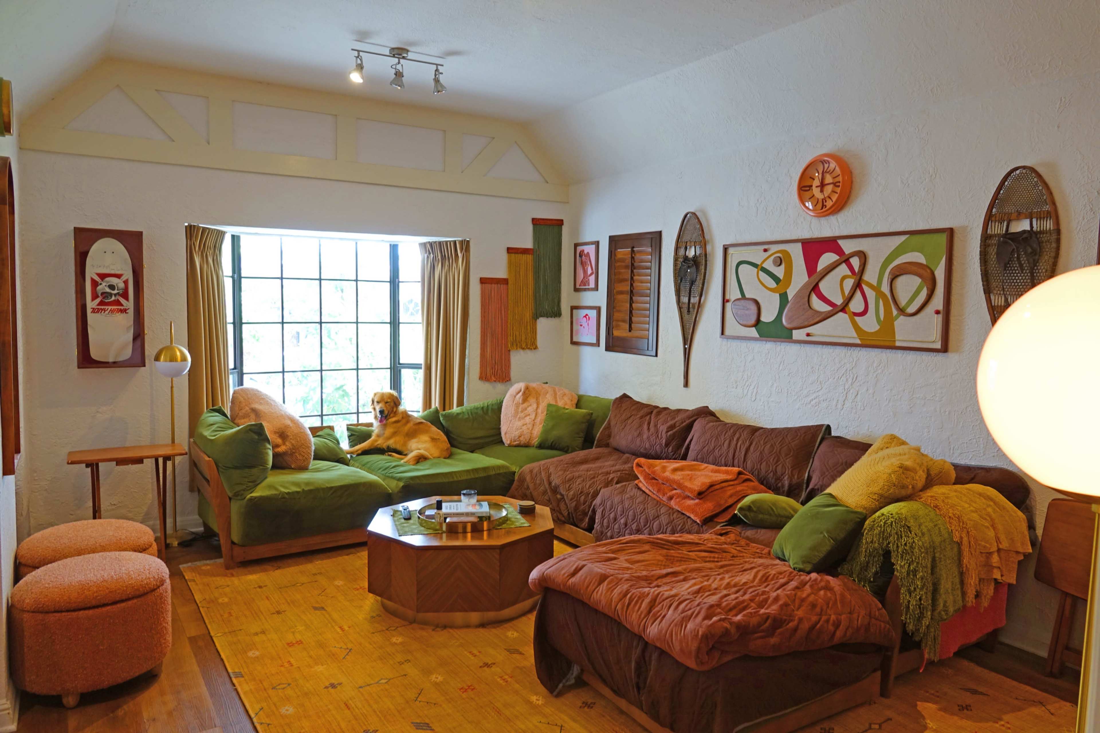 A cozy living room features green and brown sofas arranged around a wooden coffee table, with a large window allowing natural light and a dog resting on one of the sofas.
