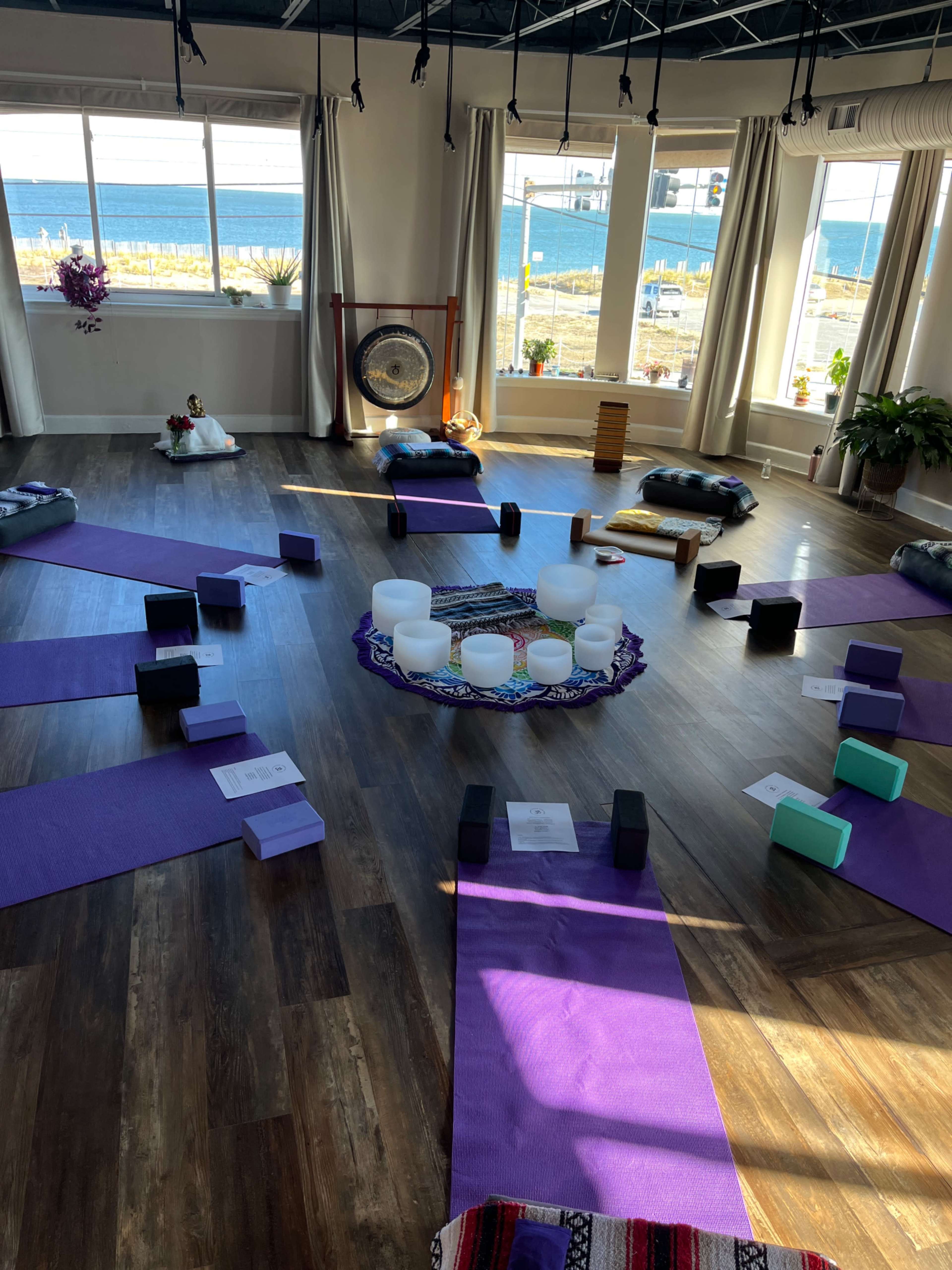 A spacious yoga studio with mats arranged in a circular formation around a center area containing singing bowls and blocks, with large windows showcasing a waterfront view.