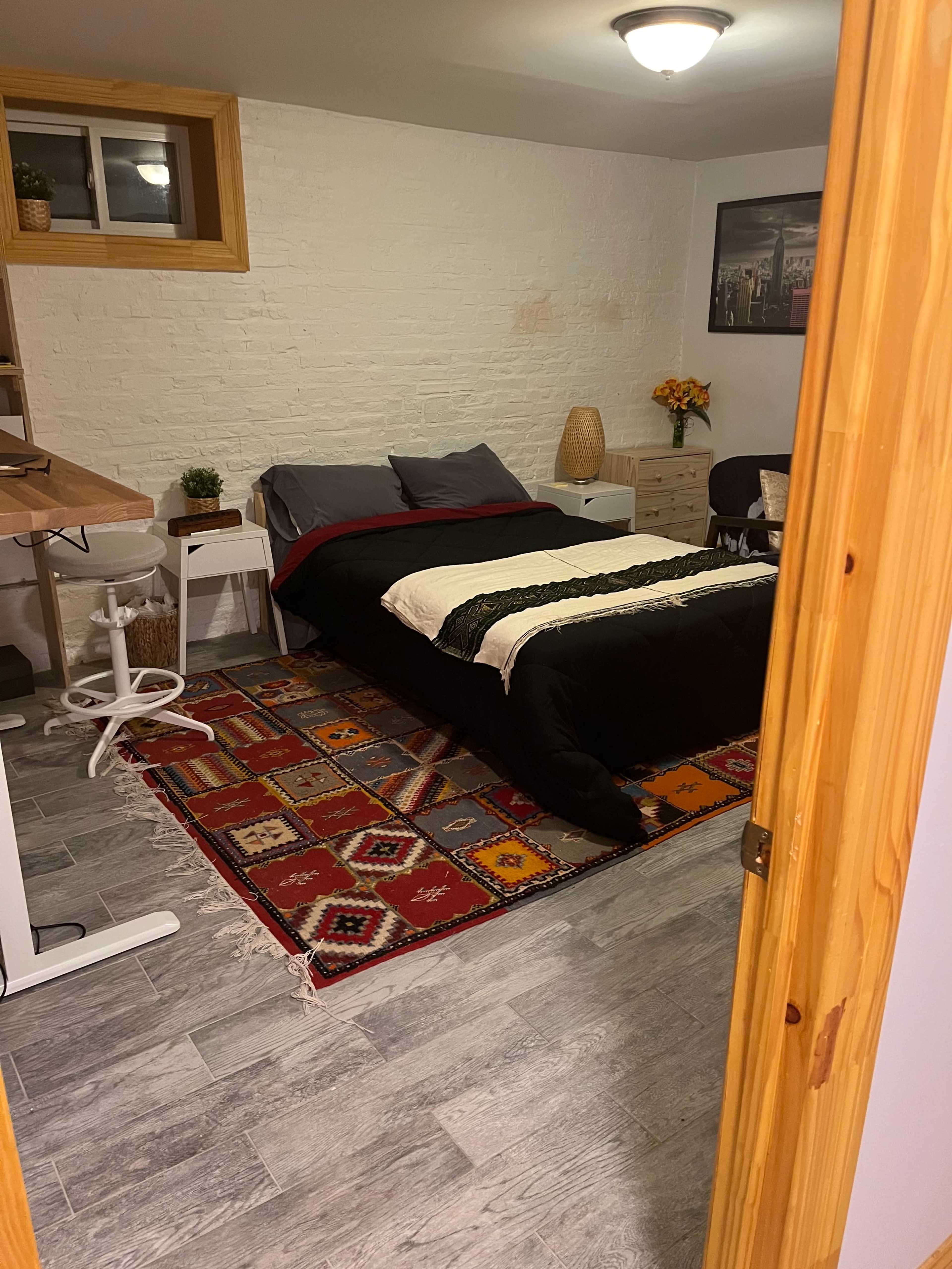 A bedroom features a bed with a black and white comforter, a patterned area rug, a small desk, and wall decor.