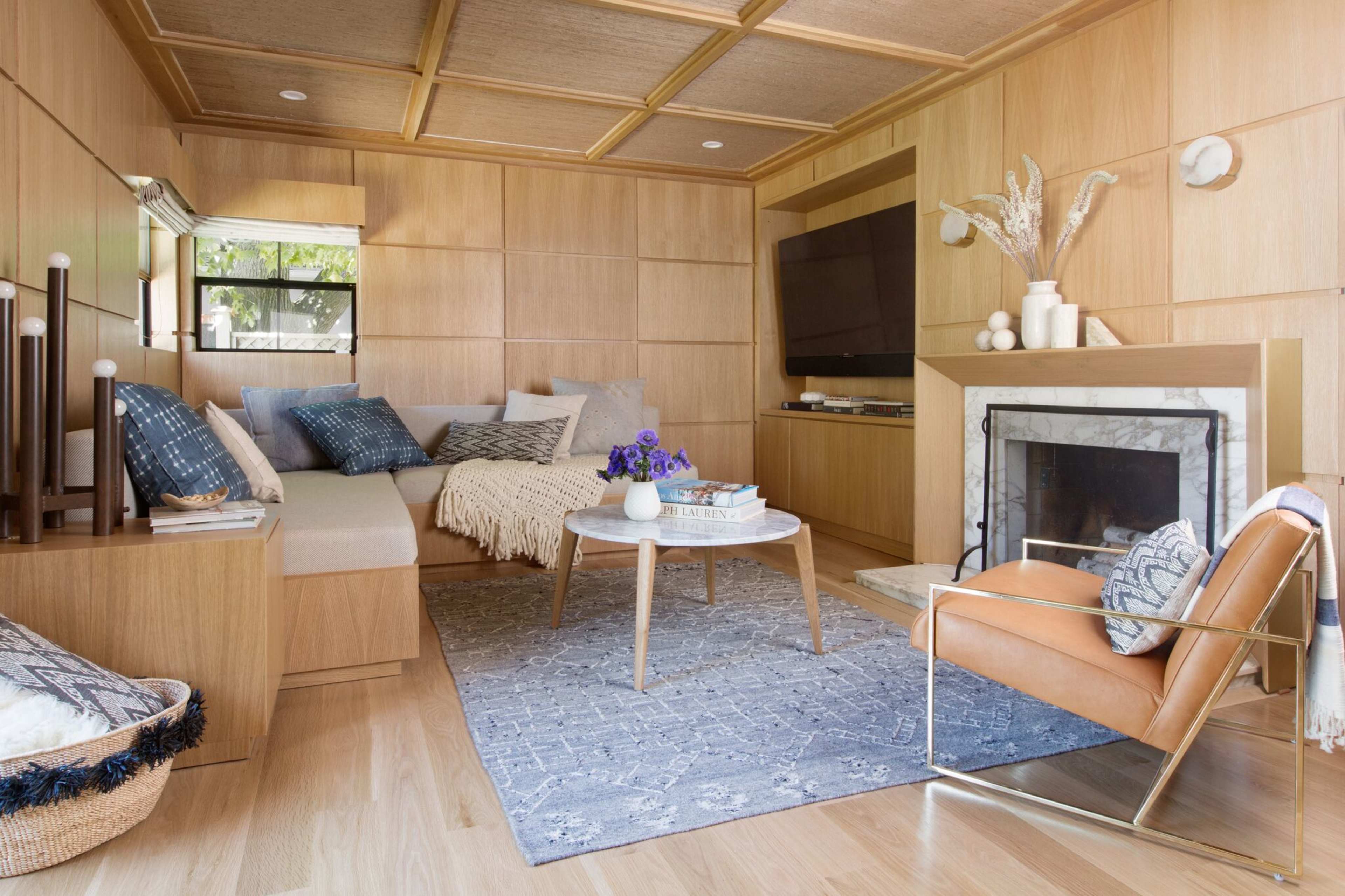 A cozy living room with wooden paneling, a marble fireplace, a coffee table, and a seating area featuring a light-colored sofa and an orange armchair.