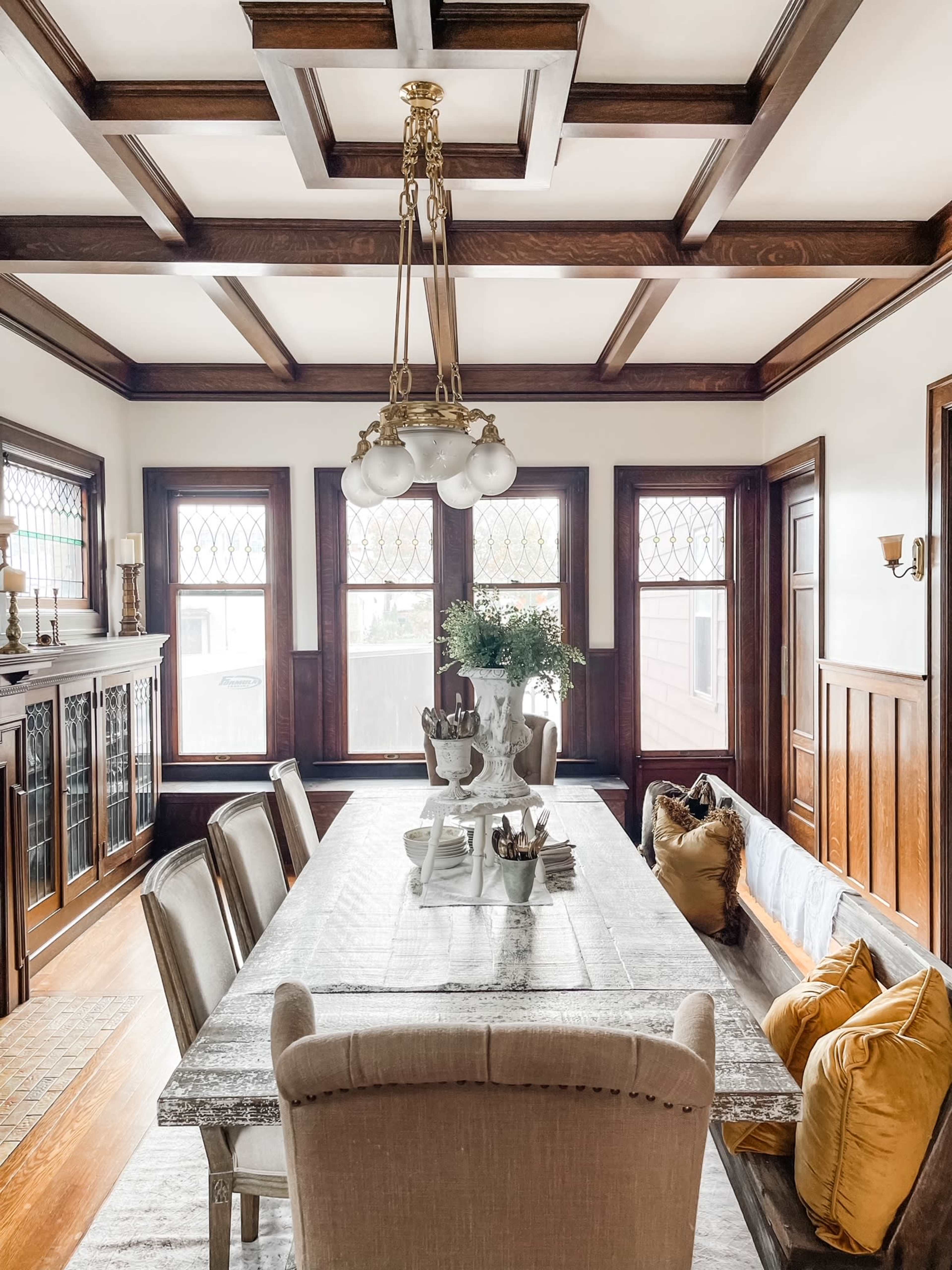 A wooden dining room features a long table set with decorative items and a chandelier, surrounded by upholstered chairs beneath a coffered ceiling with large windows.
