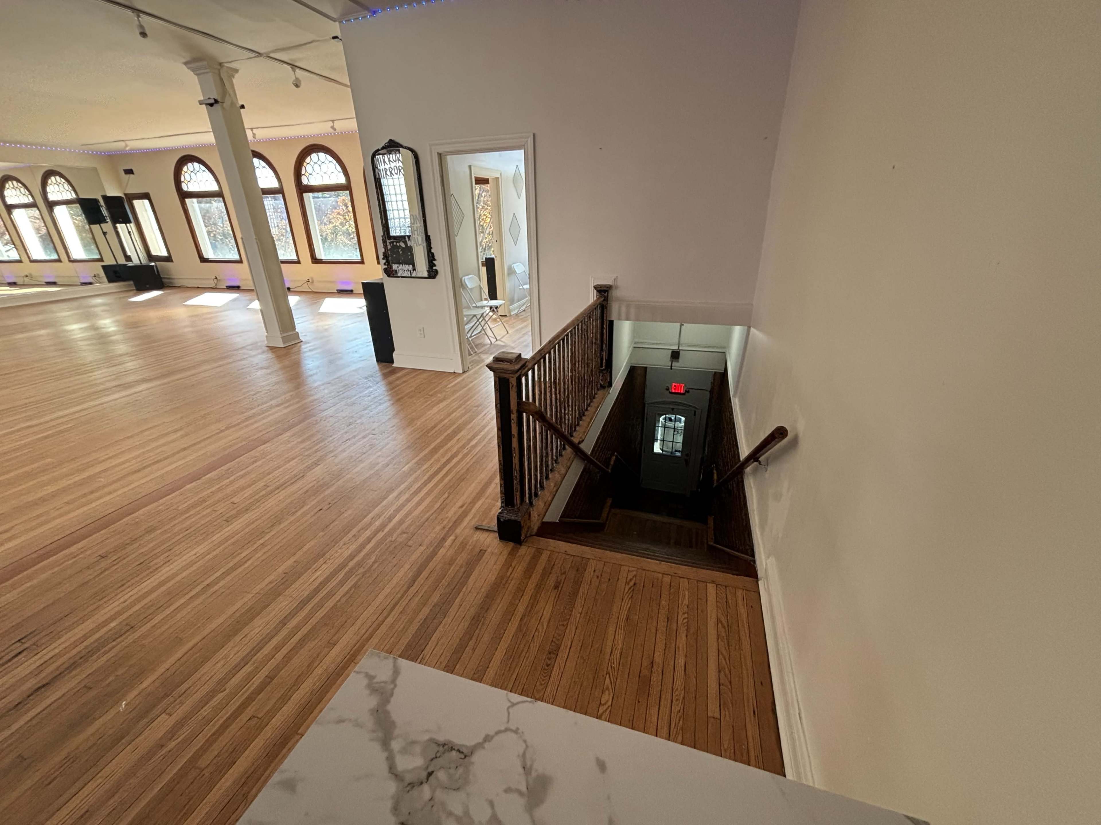 The image shows a staircase leading down to a lower level in a spacious, light-filled room with wooden flooring and large arched windows.
