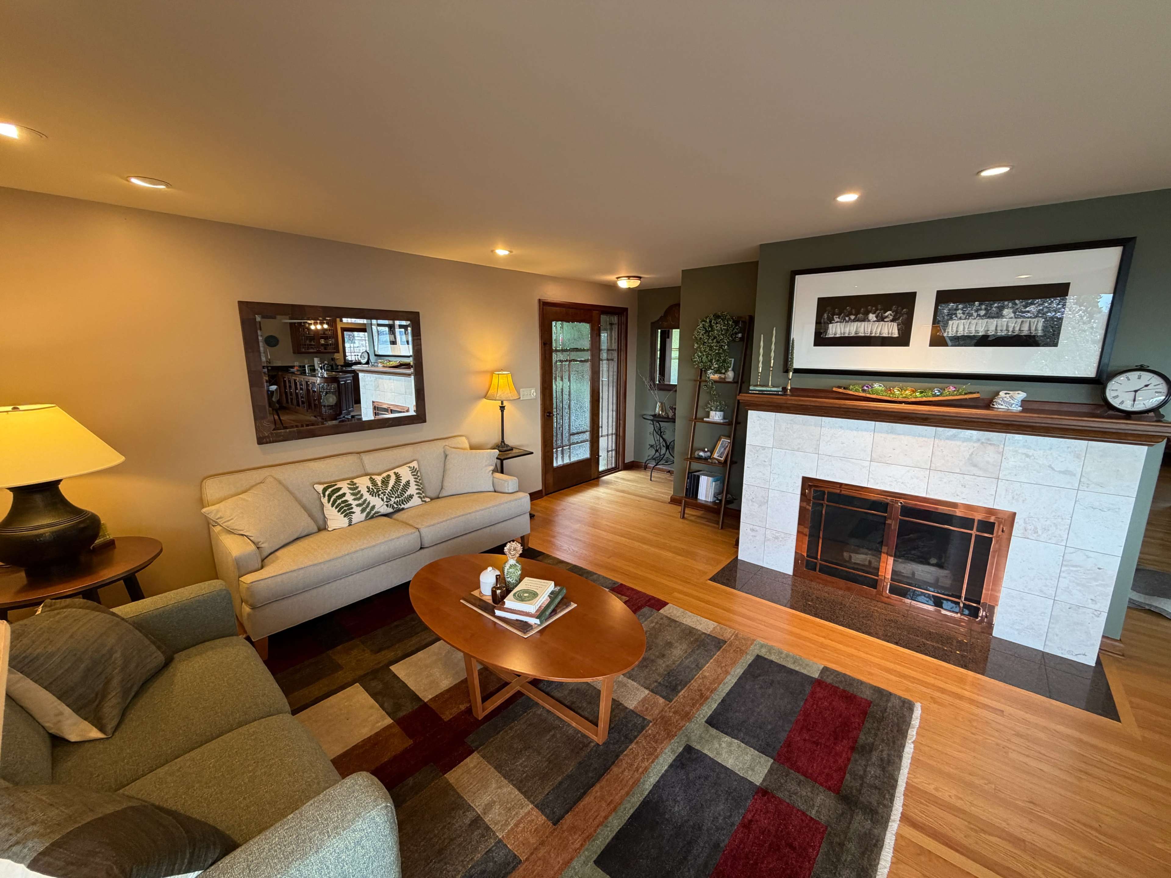 A cozy living room features a sofa, an oval coffee table, a fireplace with decorative tiles, and hardwood flooring.