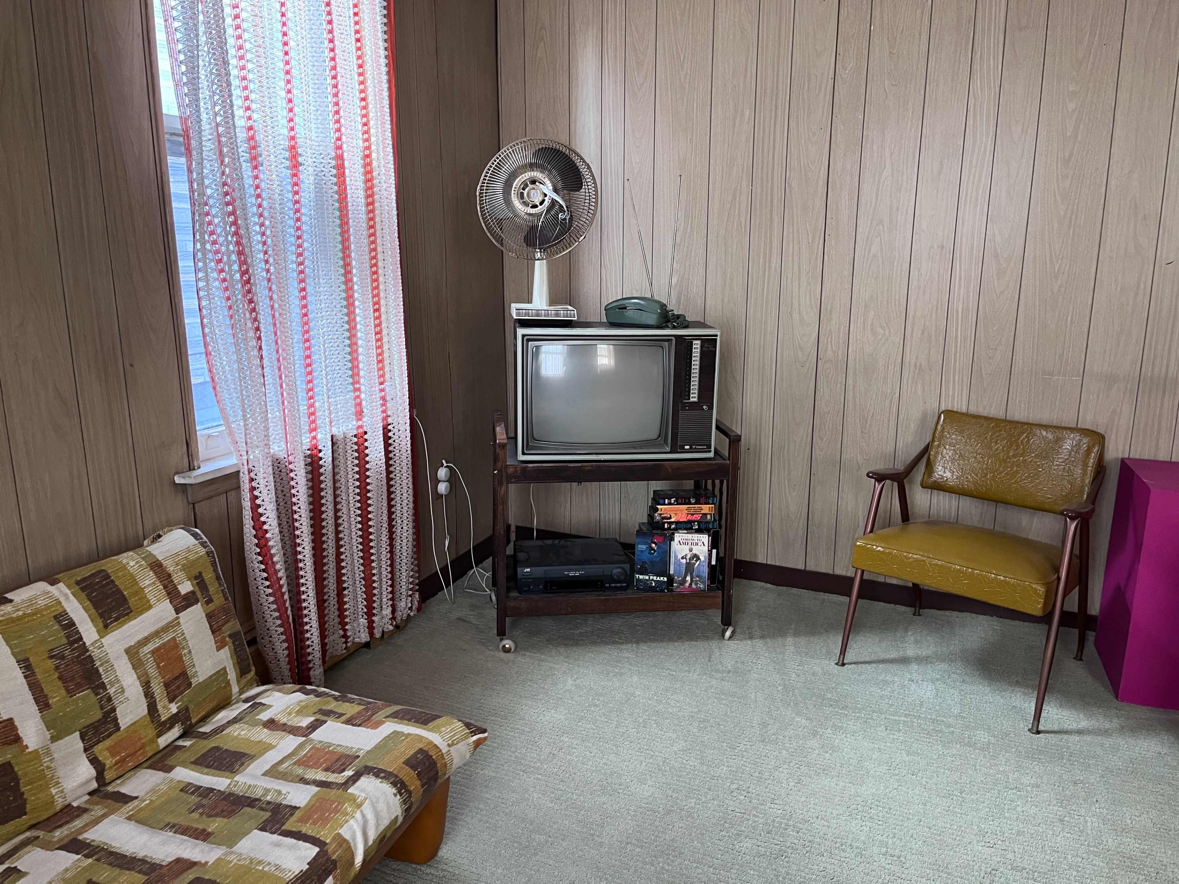 The image shows a vintage living room with a patterned sofa, a retro television on a stand, a fan, an armchair, and a window with lace curtains.