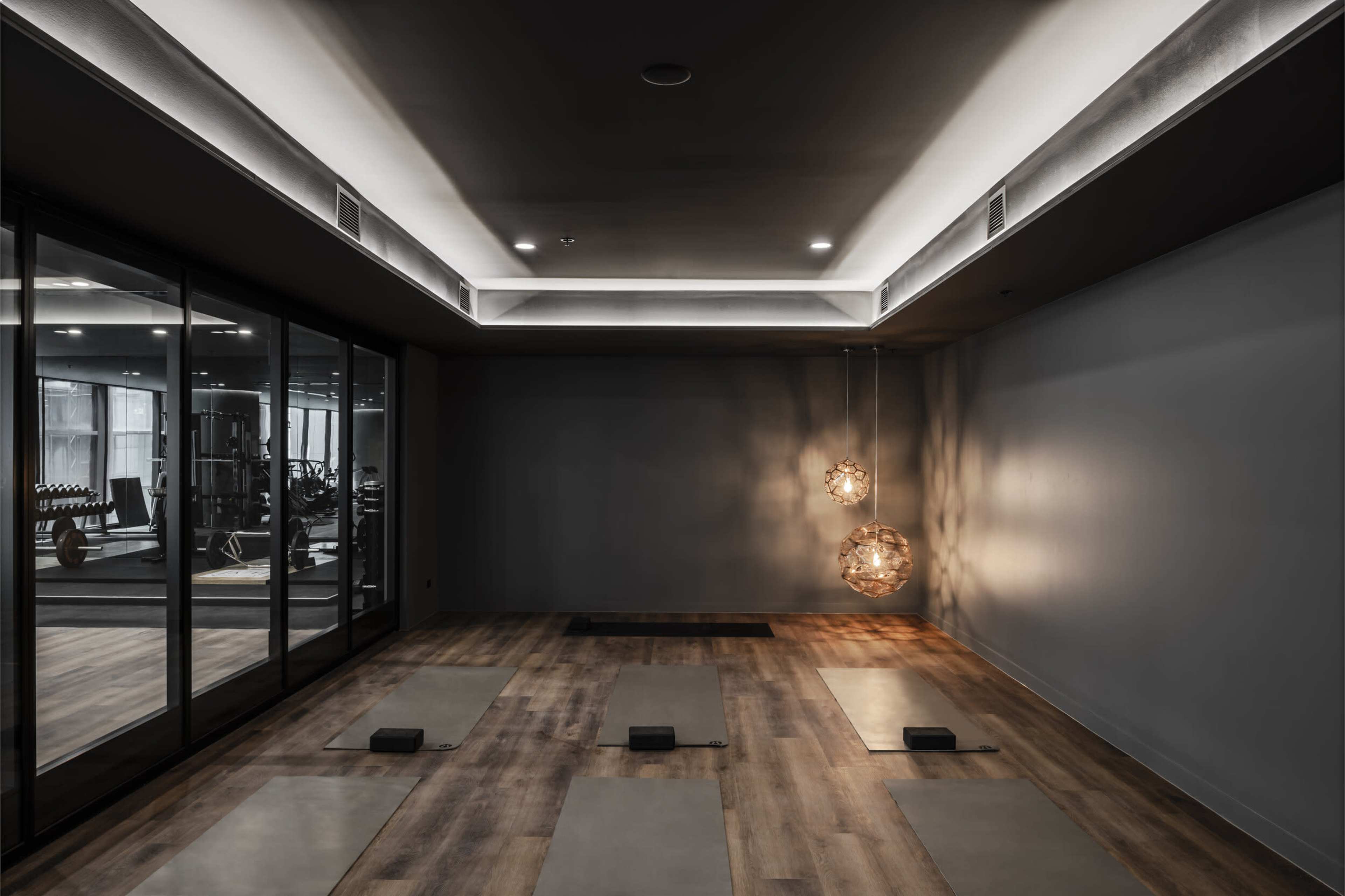 A dimly lit yoga studio features two hanging light fixtures and several yoga mats aligned on a wooden floor, with large windows revealing a gym area in the background.